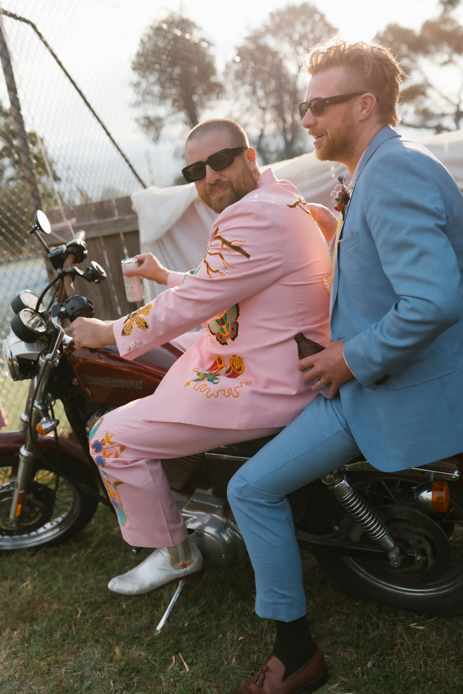 Two men in colorful suits sitting on a motorcycle outdoors, one in pink floral suit, one in blue blazer, both wearing sunglasses.