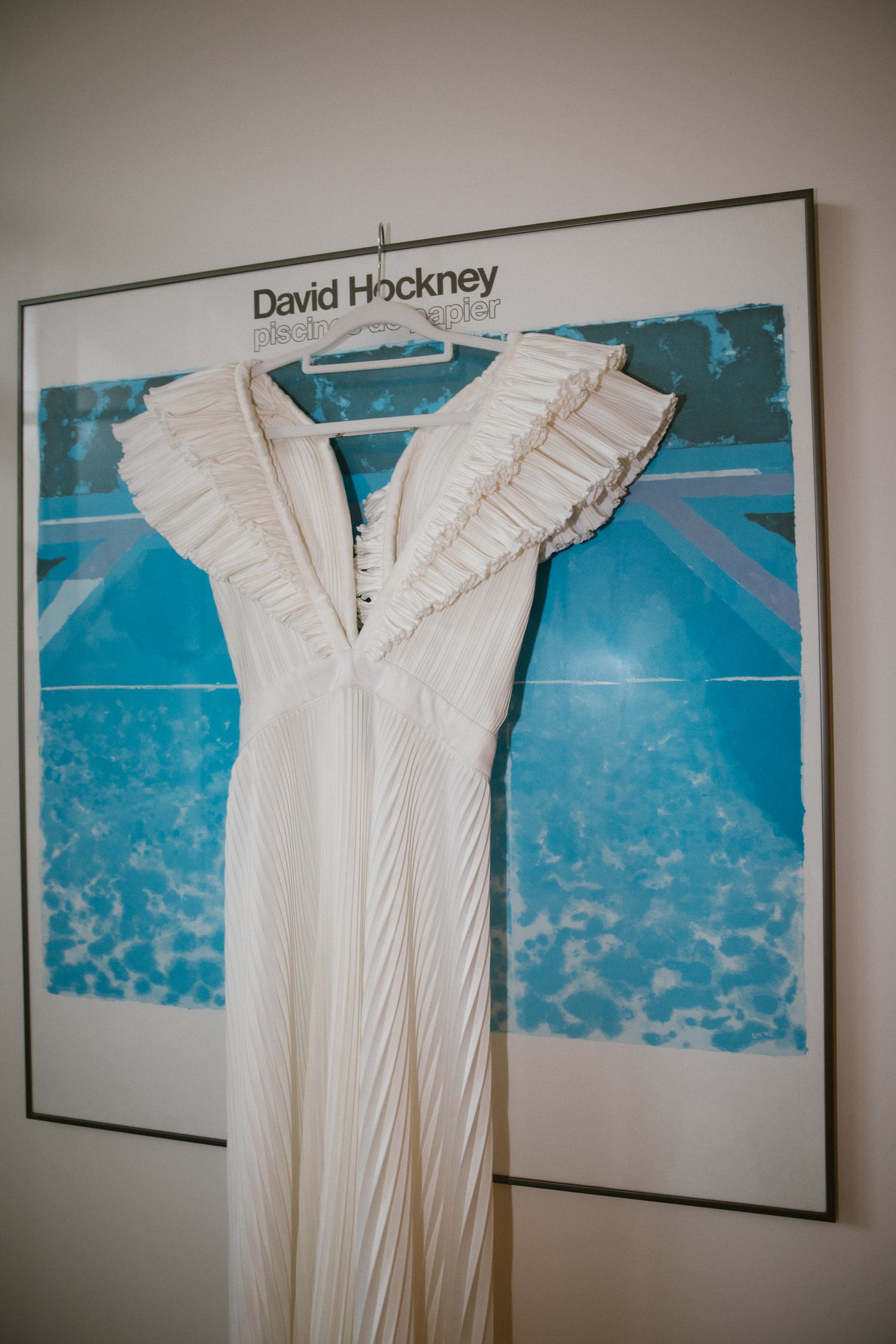 White pleated wedding dress with ruffle shoulders hanging on a David Hockney art print.
