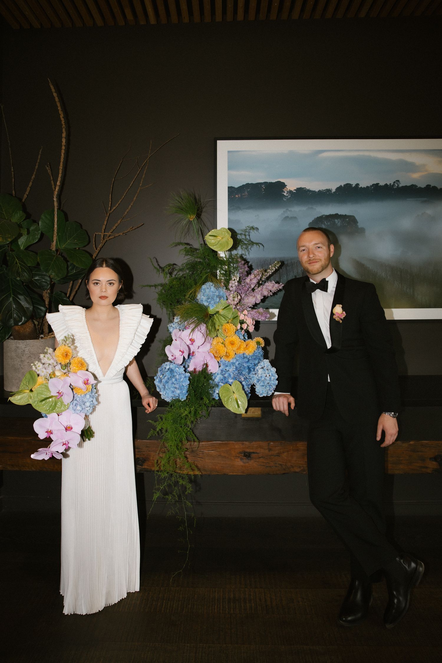Bride and groom posing beside colorful floral arrangement with orchids and hydrangeas in dark reception venue.