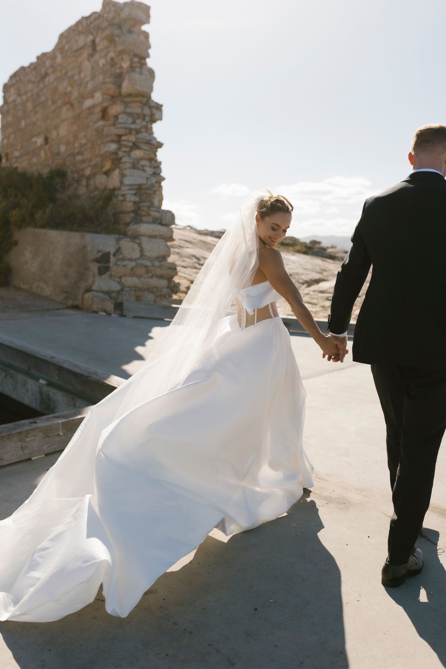Bride in flowing white gown and veil holding grooms hand near stone ruins.