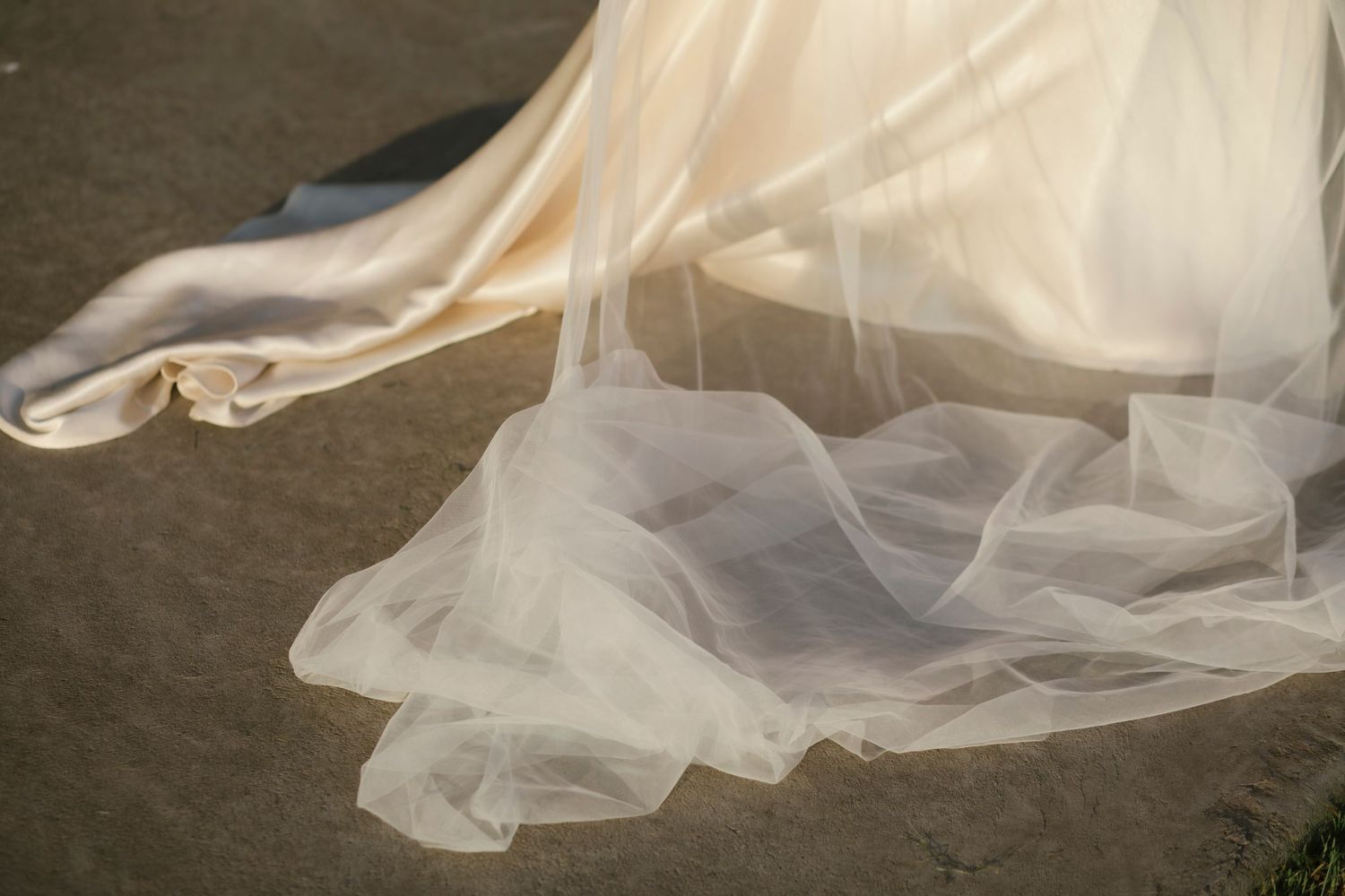 Close-up of a flowing ivory satin wedding dress train with delicate tulle overlay on pavement.