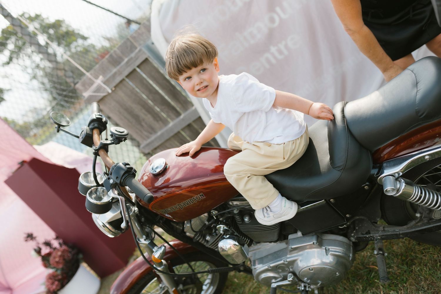 Young boy smiling while sitting on a red motorcycle outdoors at a casual event.
