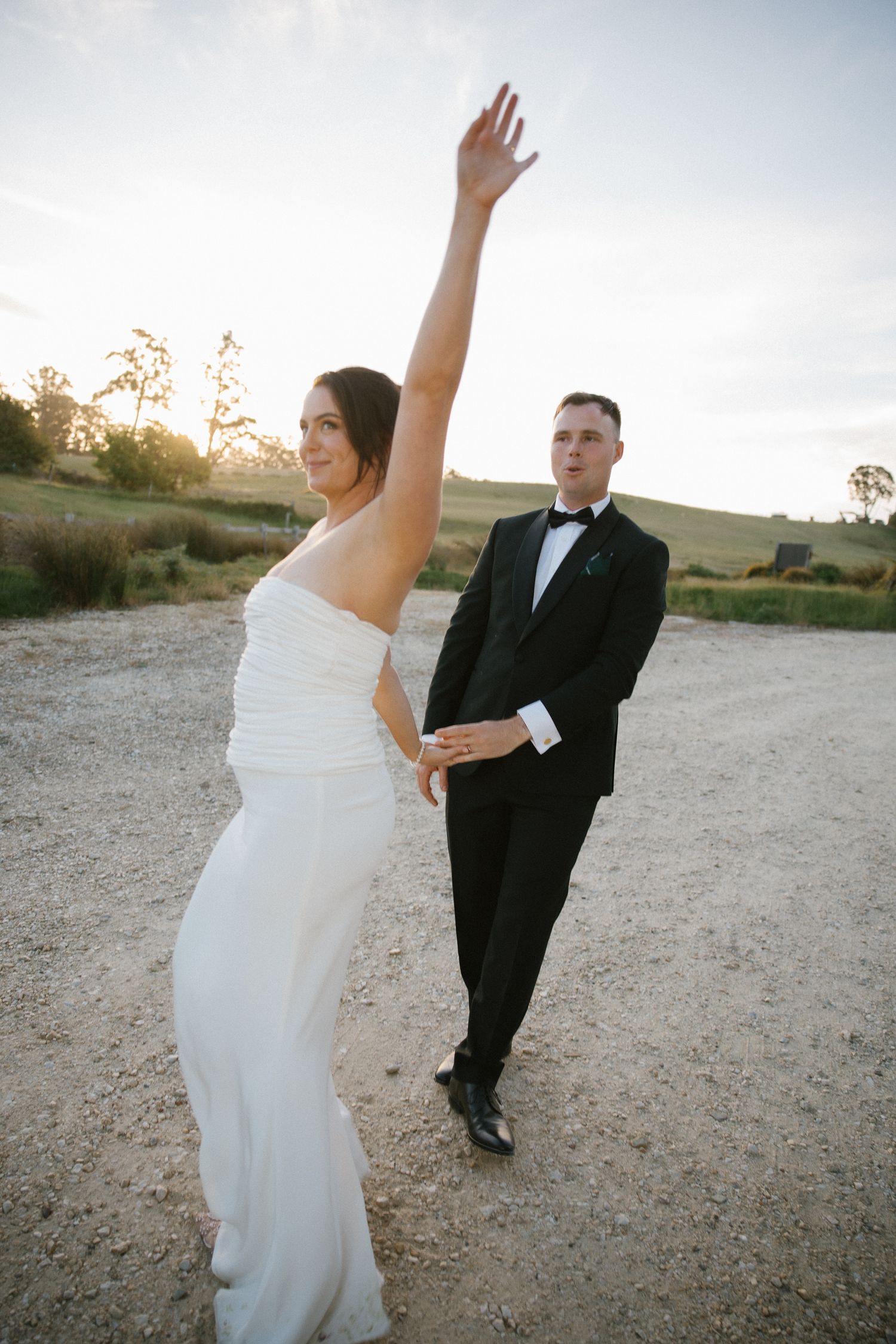 Joyful bride raising arm while groom watches during sunset golden hour wedding portraits.