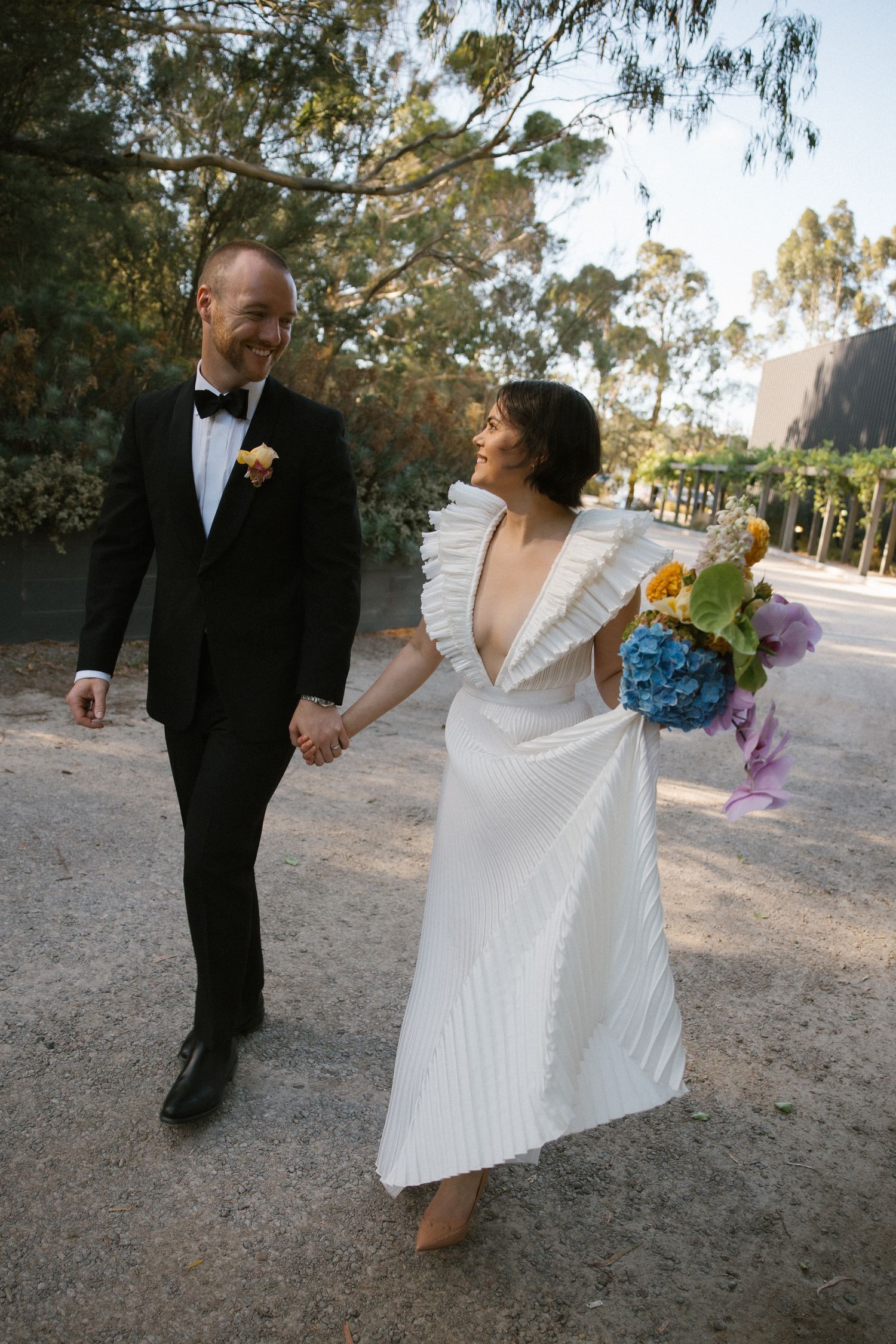 Happy bride and groom walking hand in hand outdoors, bride carrying colorful wedding bouquet.