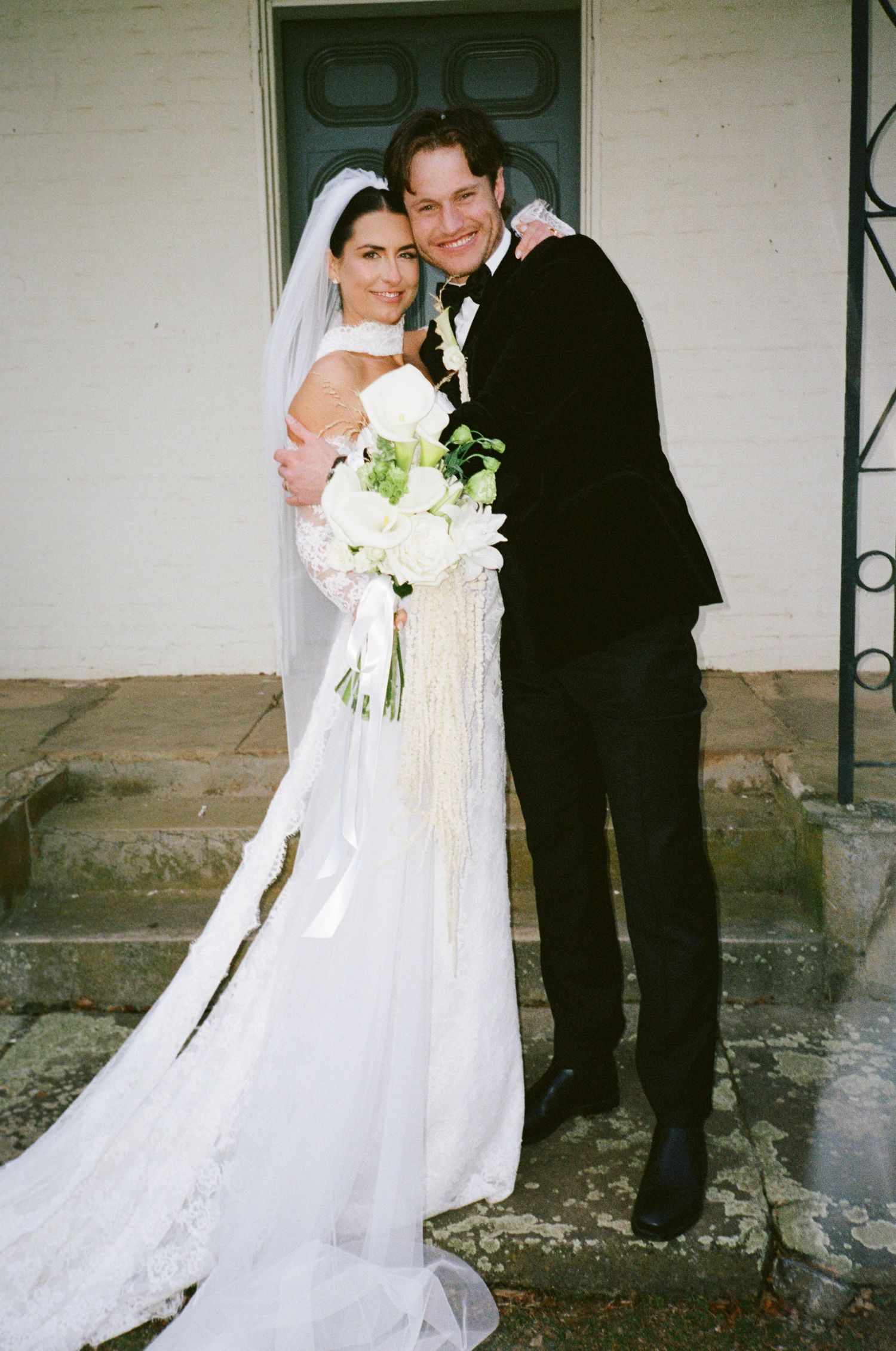 Bride in white gown with veil holding bouquet stands with groom in black tuxedo on church steps.