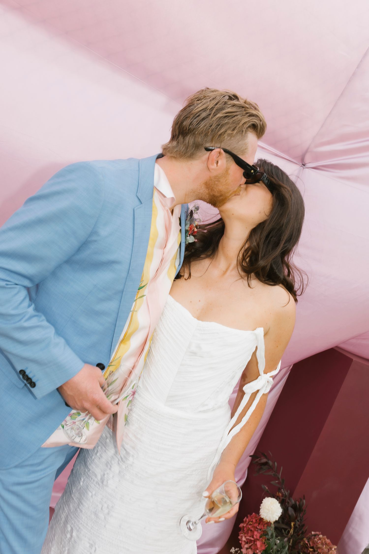 Bride and groom kissing near pink bouncy castle, groom in blue suit, bride in white dress.