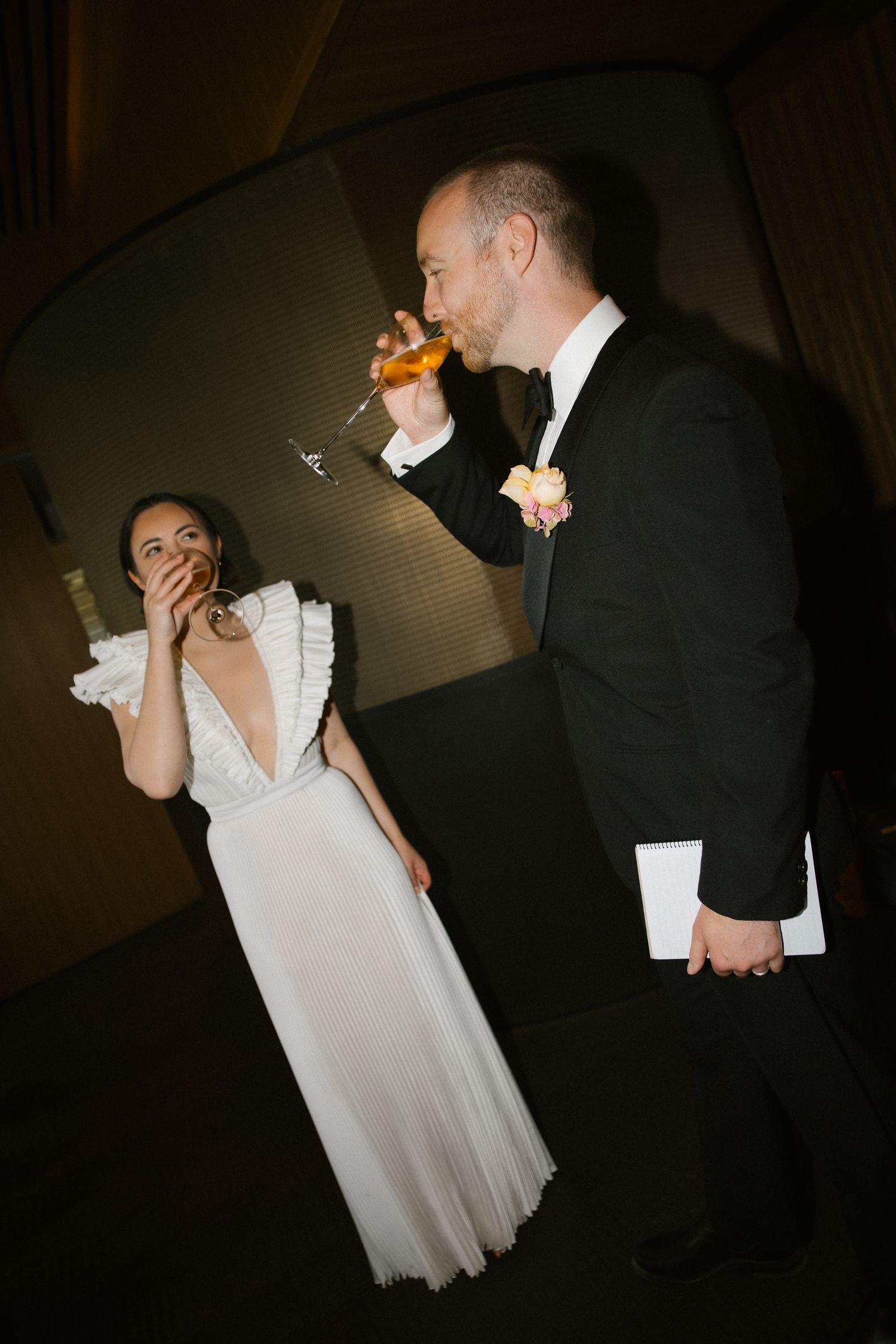 Bride laughing while groom in tuxedo drinks from glass during wedding reception in dark venue.