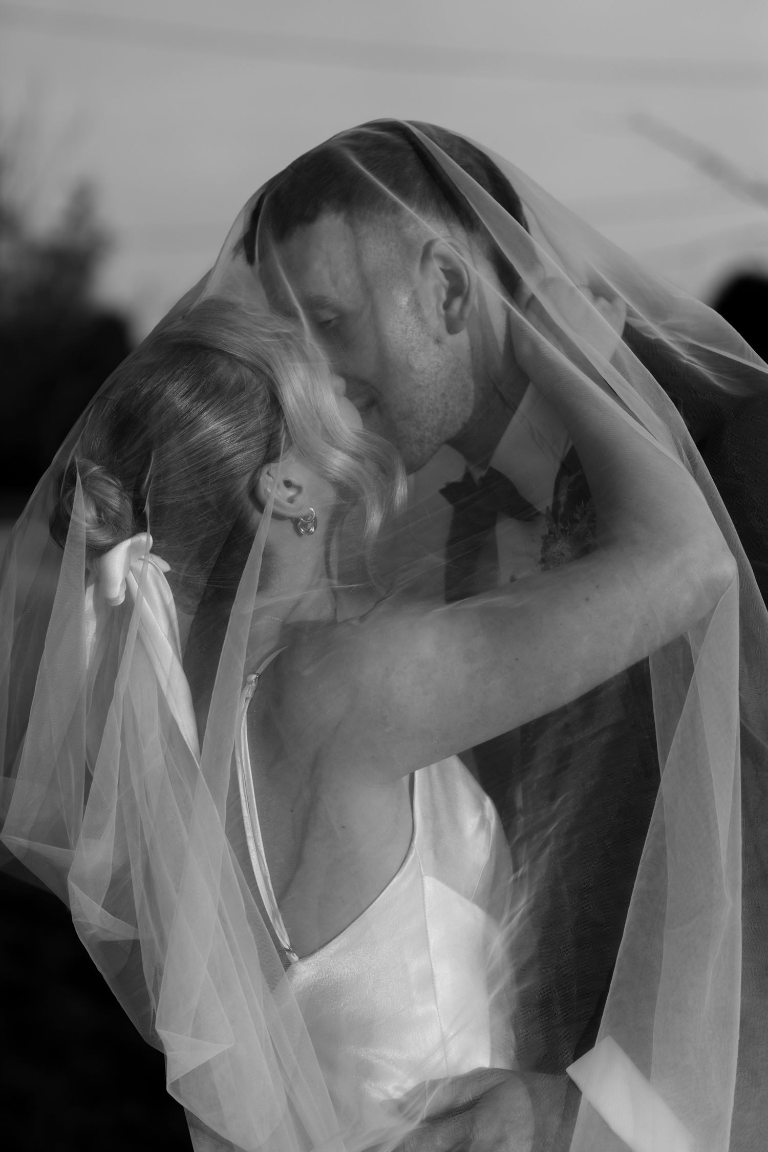 Black and white romantic photo of bride and groom kissing under flowing wedding veil.