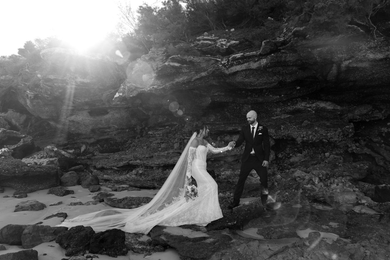 Black and white photo of bride and groom walking on rocky beach with dramatic sunflare.