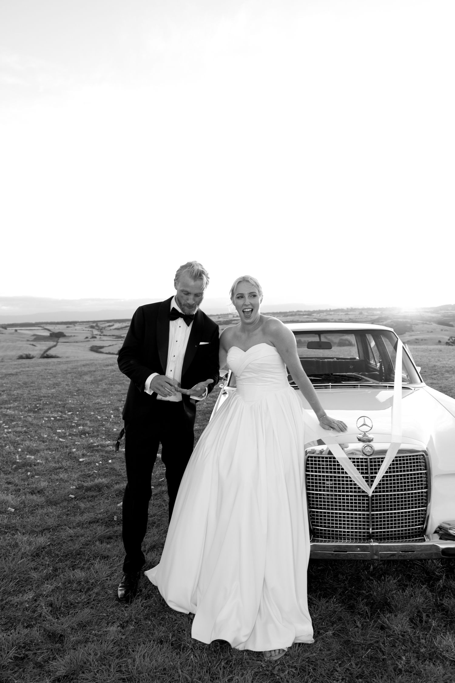 Bride and groom laughing beside a vintage Mercedes wedding car in black and white photo.