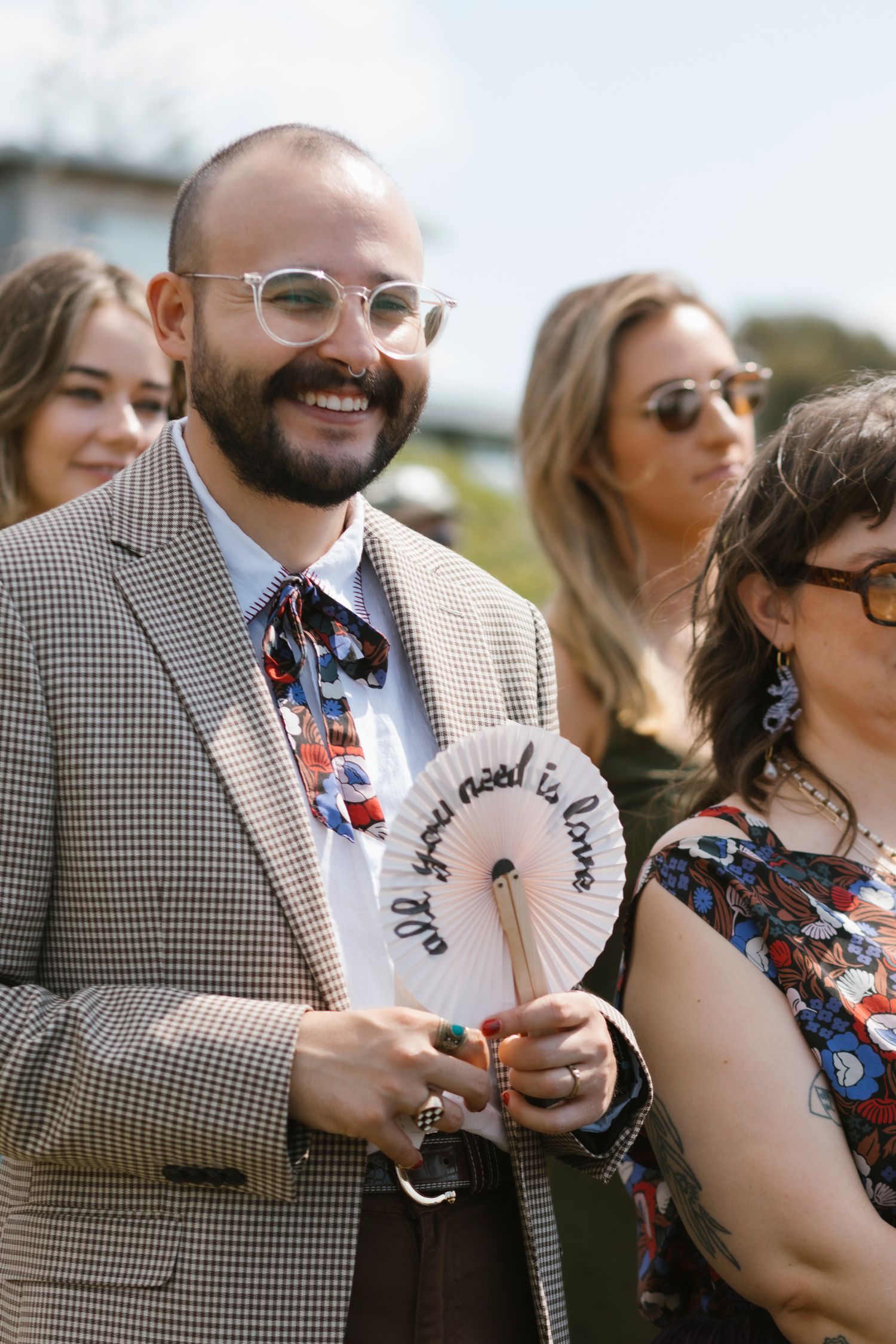 Smiling bearded wedding guest in checkered suit holding decorative fan reading all you need is love.
