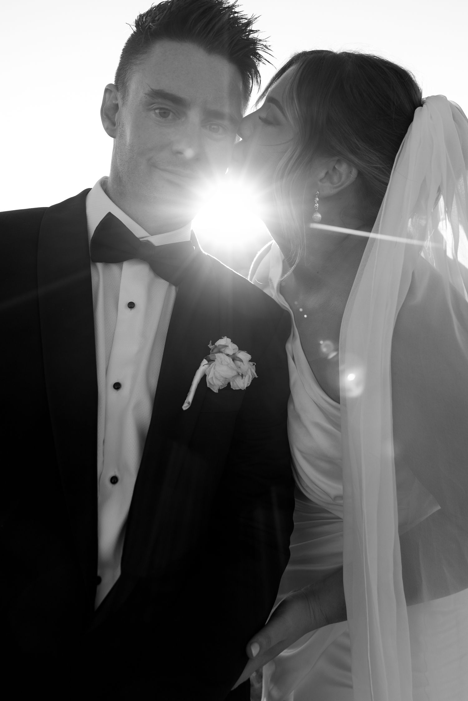 Bride kissing groom on the cheek with a sunburst lens flare effect in a classic black and white wedding portrait.