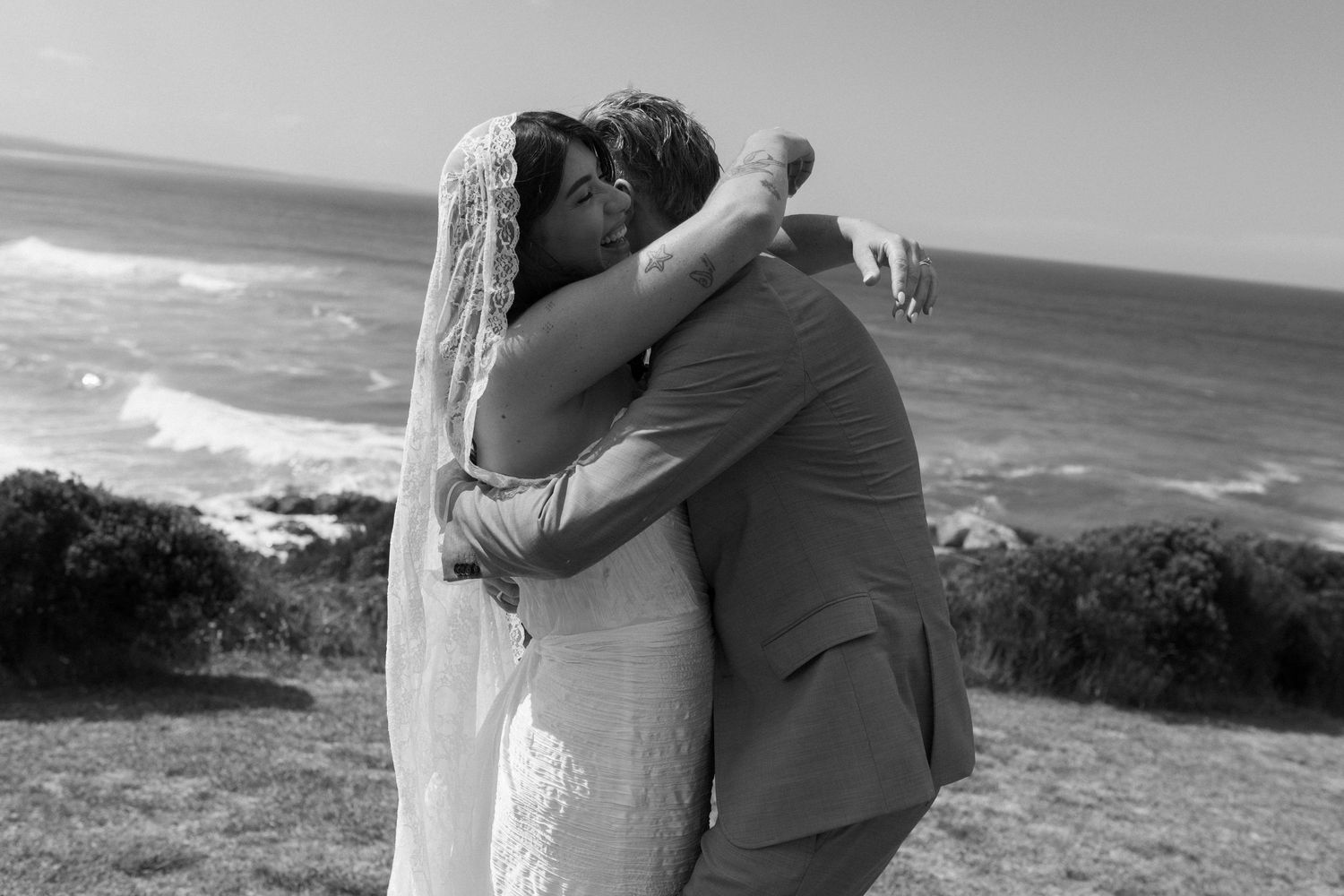 Black and white photo of a bride and groom embracing on a coastal cliff with ocean waves in the background.