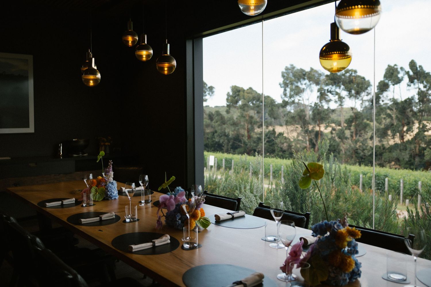 Elegant winery dining room with long wooden table, colorful floral centerpieces, and vineyard views.