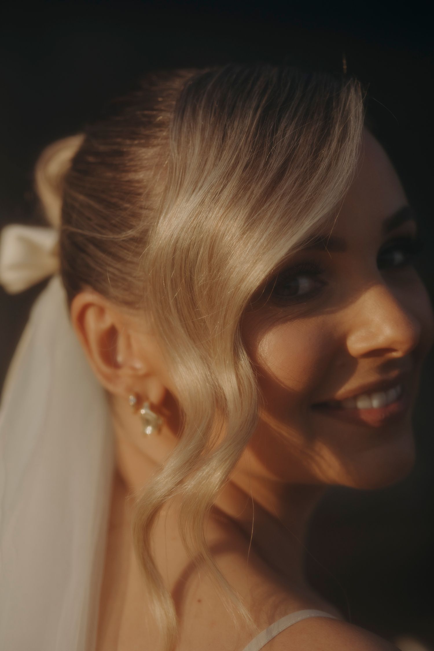 Smiling blonde bride with soft waves, pearl earrings and white bow veil in golden light.