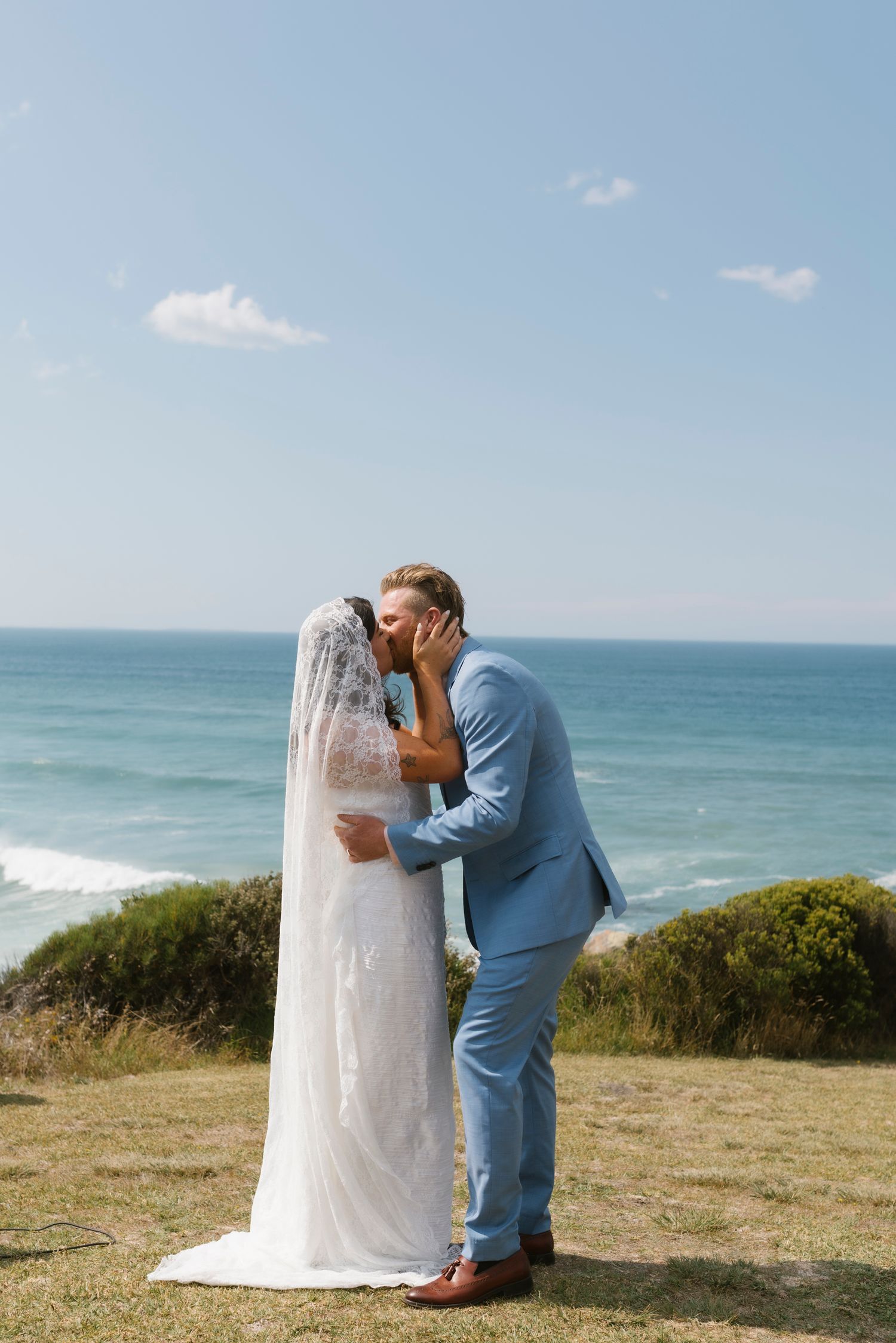 Bride and groom share a kiss at a scenic coastal wedding venue with ocean views and blue sky.