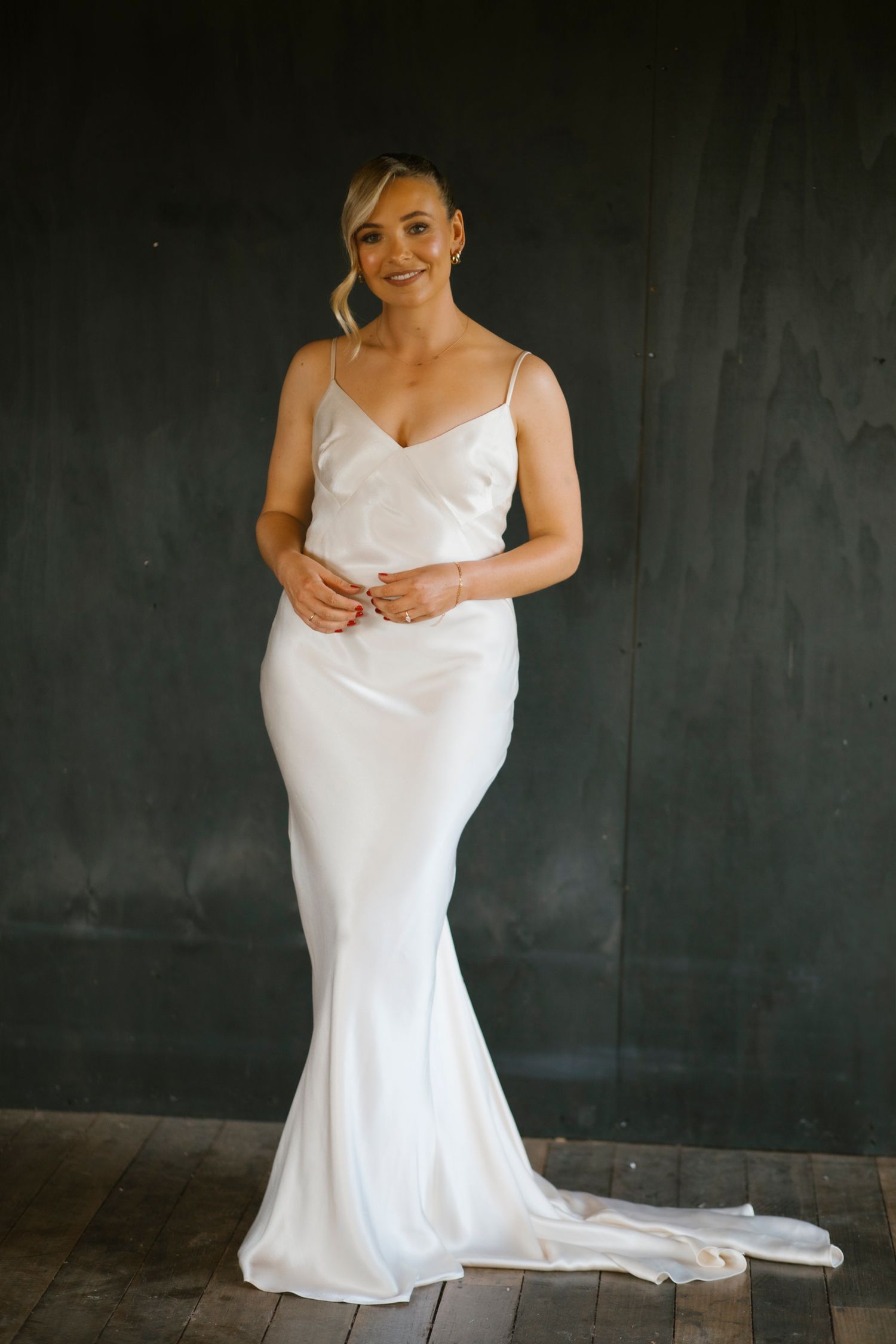 Elegant blonde bride posing in a fitted white satin slip wedding dress against a dark wooden backdrop.