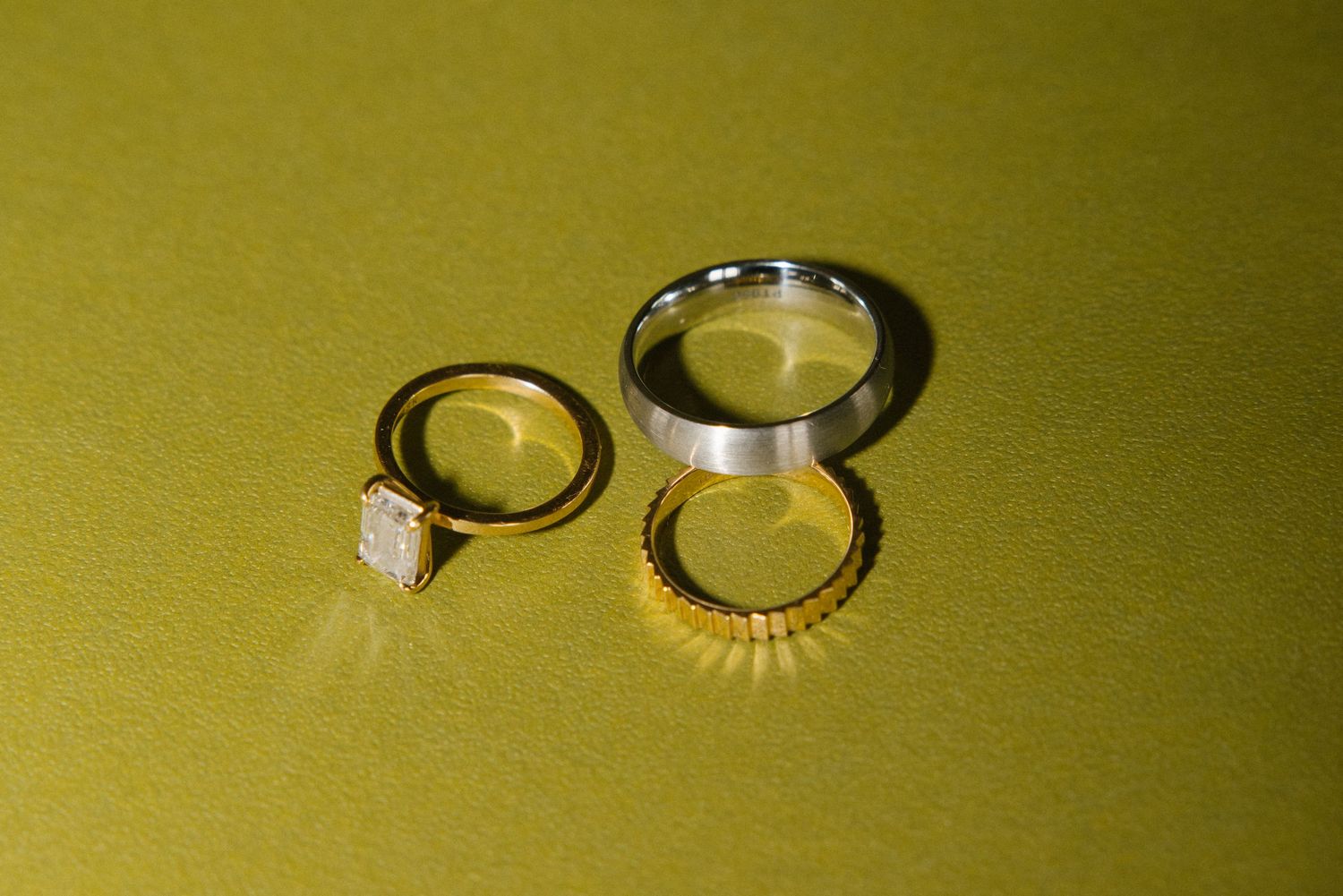 Three wedding rings on gold surface including diamond engagement ring, gold band, and silver band.