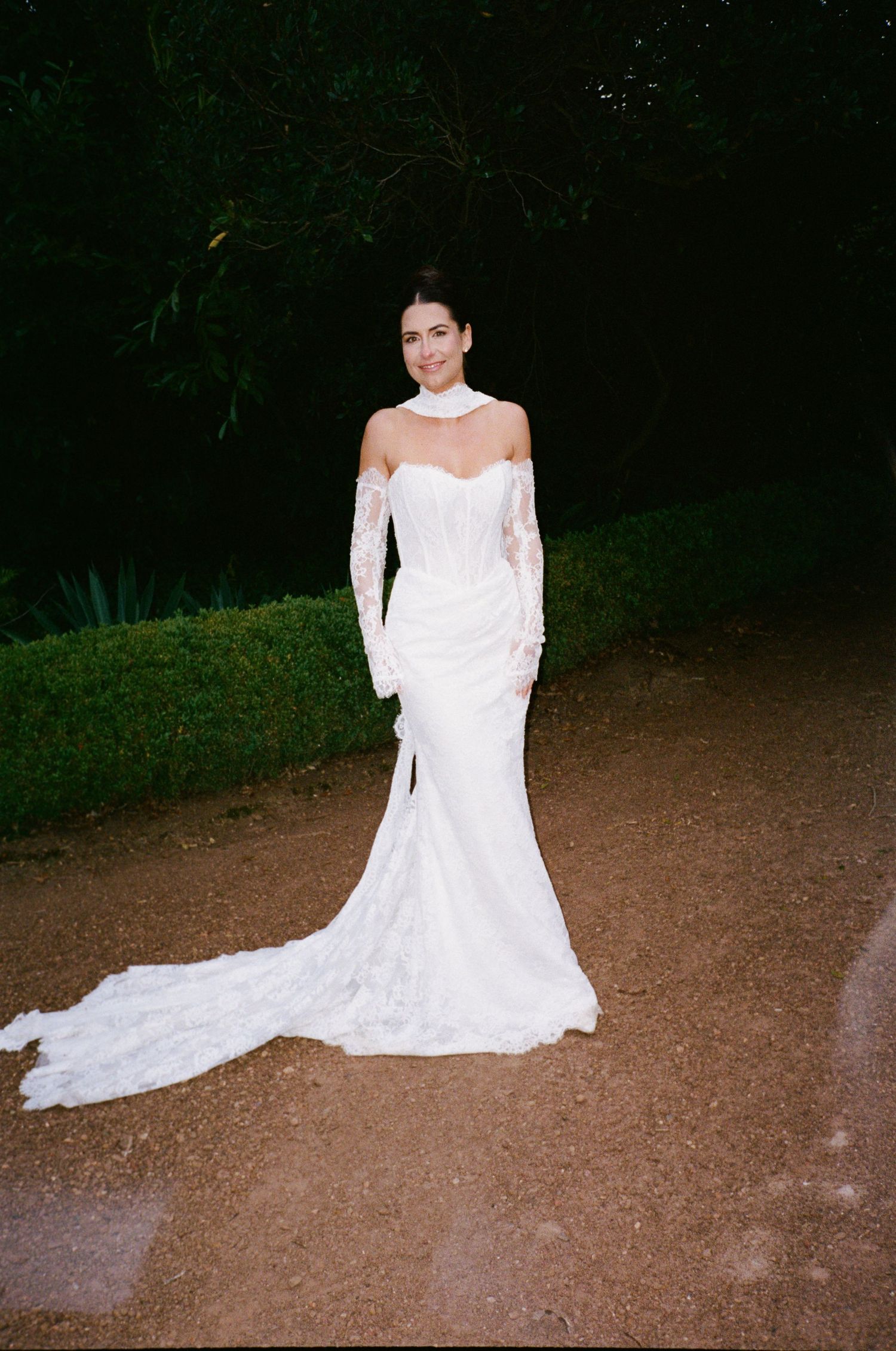 Bride in elegant white lace off-shoulder mermaid wedding gown with long train standing on gravel path.