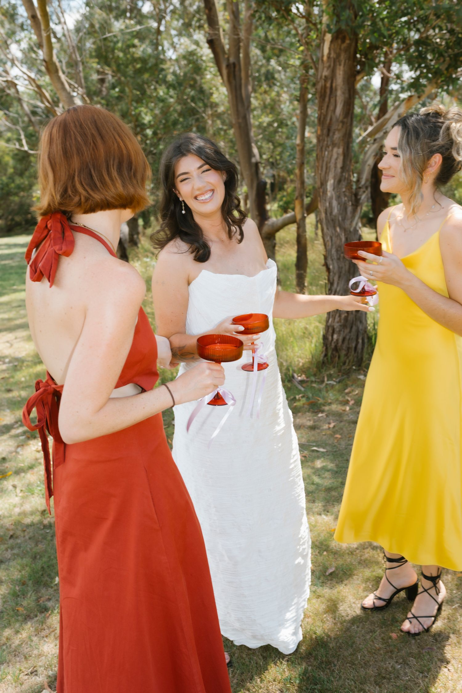 Bride in white dress toasting with bridesmaids in red and yellow dresses outdoors among trees.