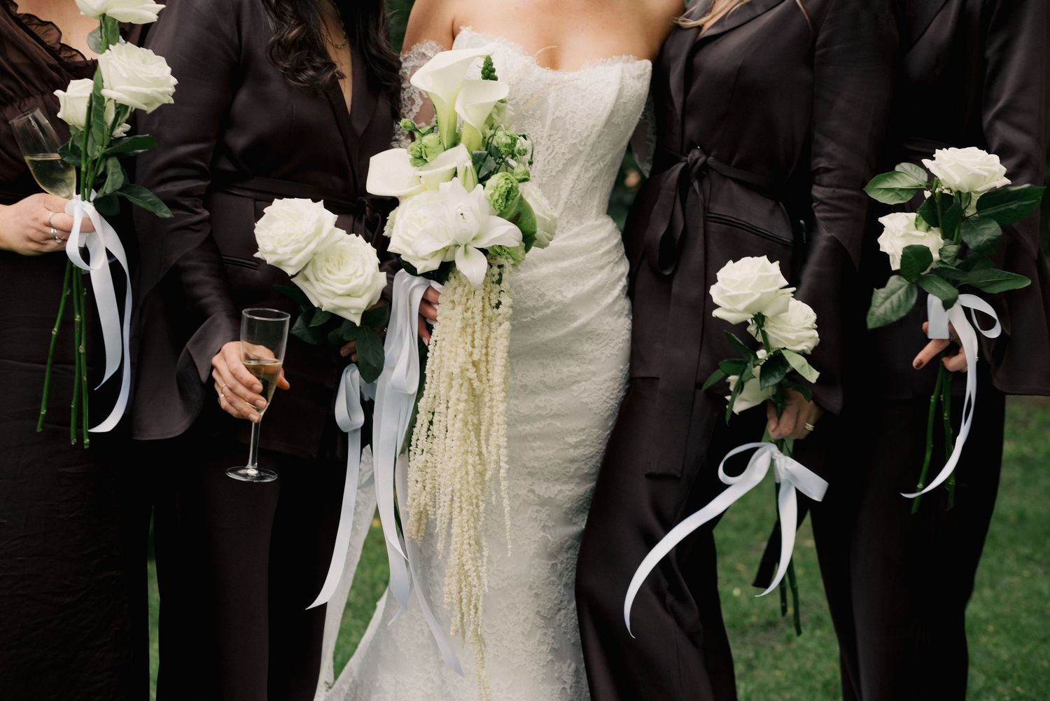 Bride in fitted white gown with bridesmaids in dark brown suits holding white rose bouquets with ribbon streamers.