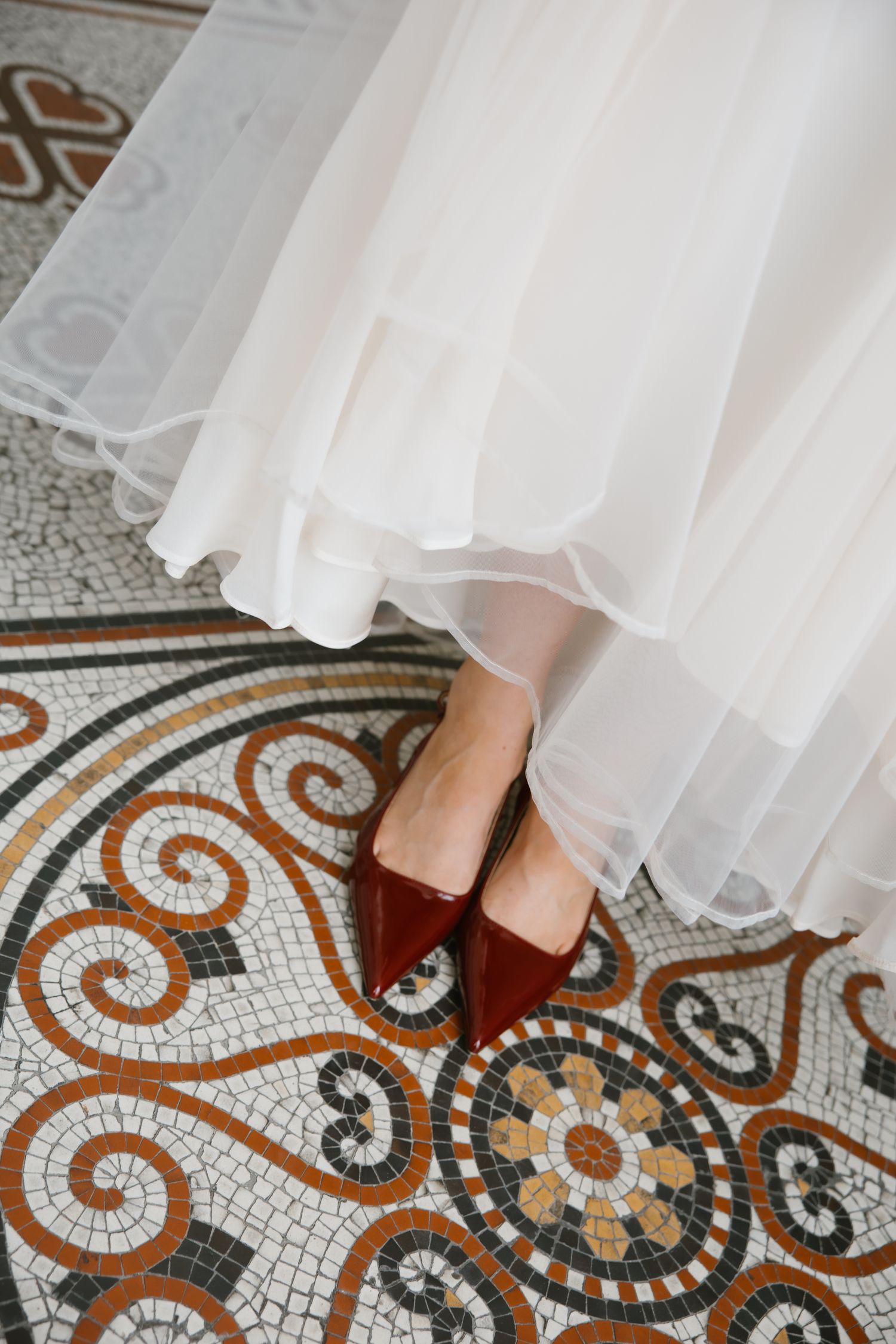 Close-up of brides feet in pointed red heels on ornate mosaic tile floor beneath white tulle wedding gown.