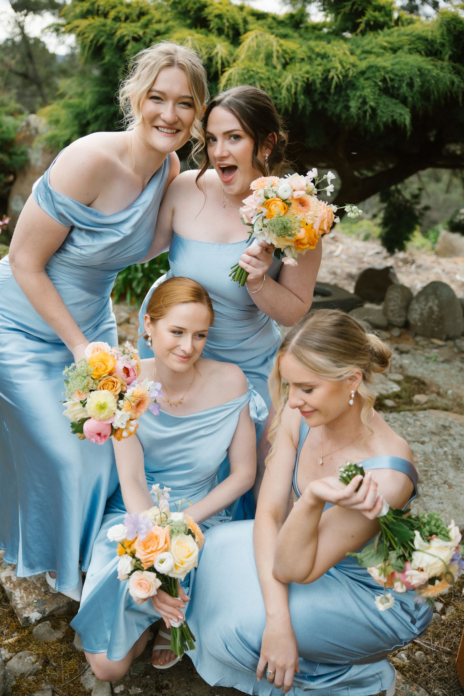 Four bridesmaids in powder blue satin dresses holding colorful bouquets posing playfully outdoors.