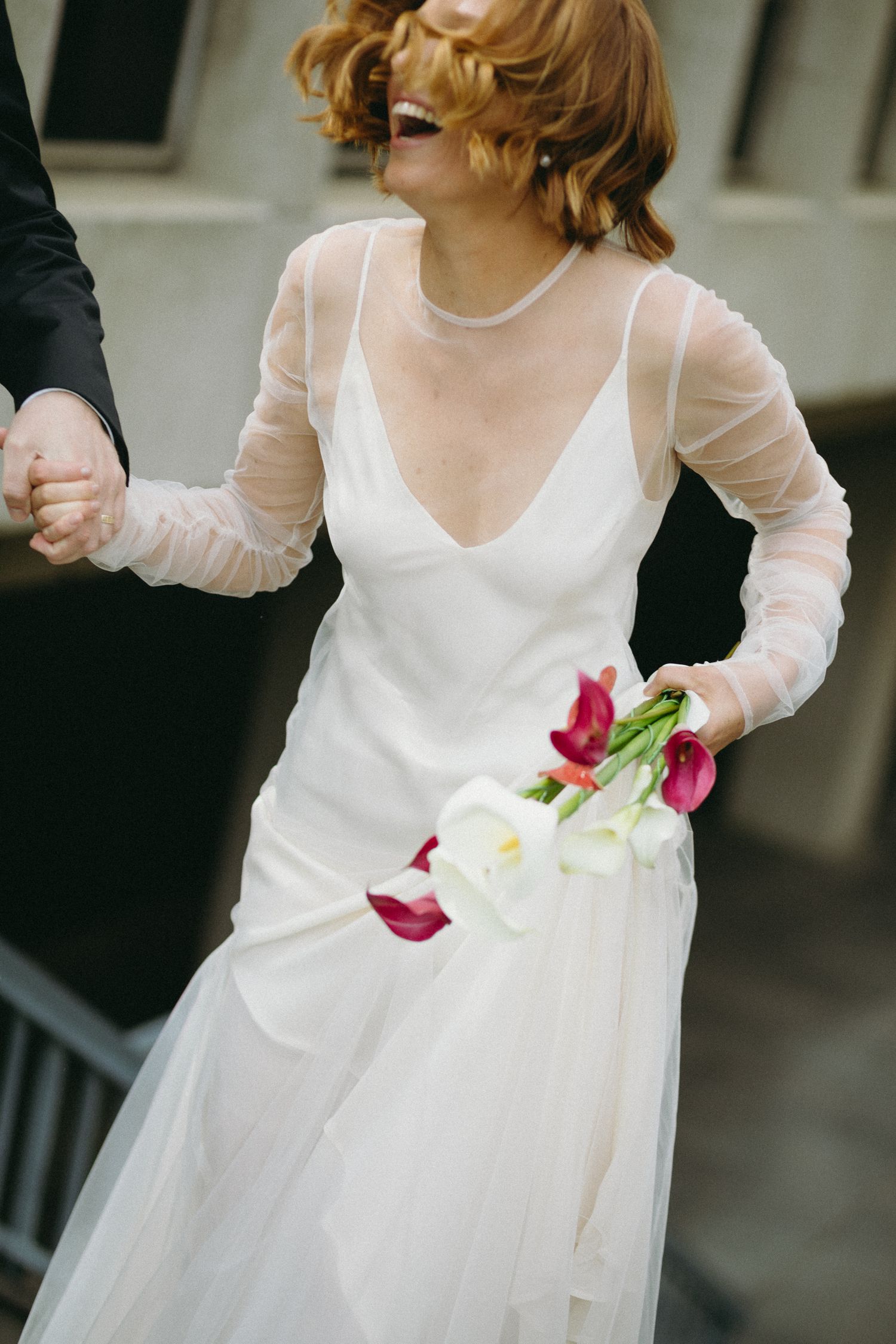 Joyful bride in white tulle gown laughing while holding hands with groom, carrying calla lilies.