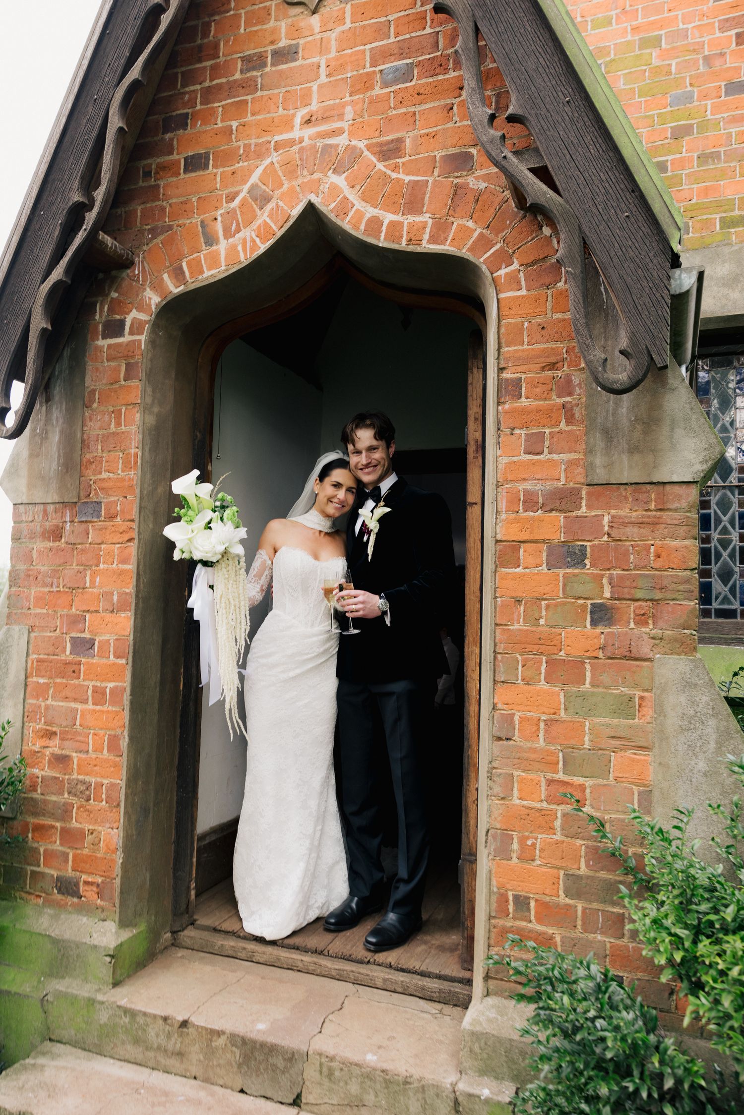 Bride and groom pose together in arched brick church doorway holding bouquet and champagne.
