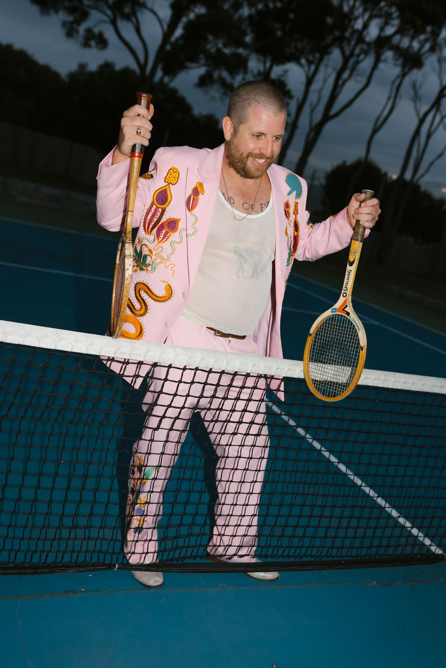 Man in a pink embroidered suit holding tennis rackets on a blue court at dusk.