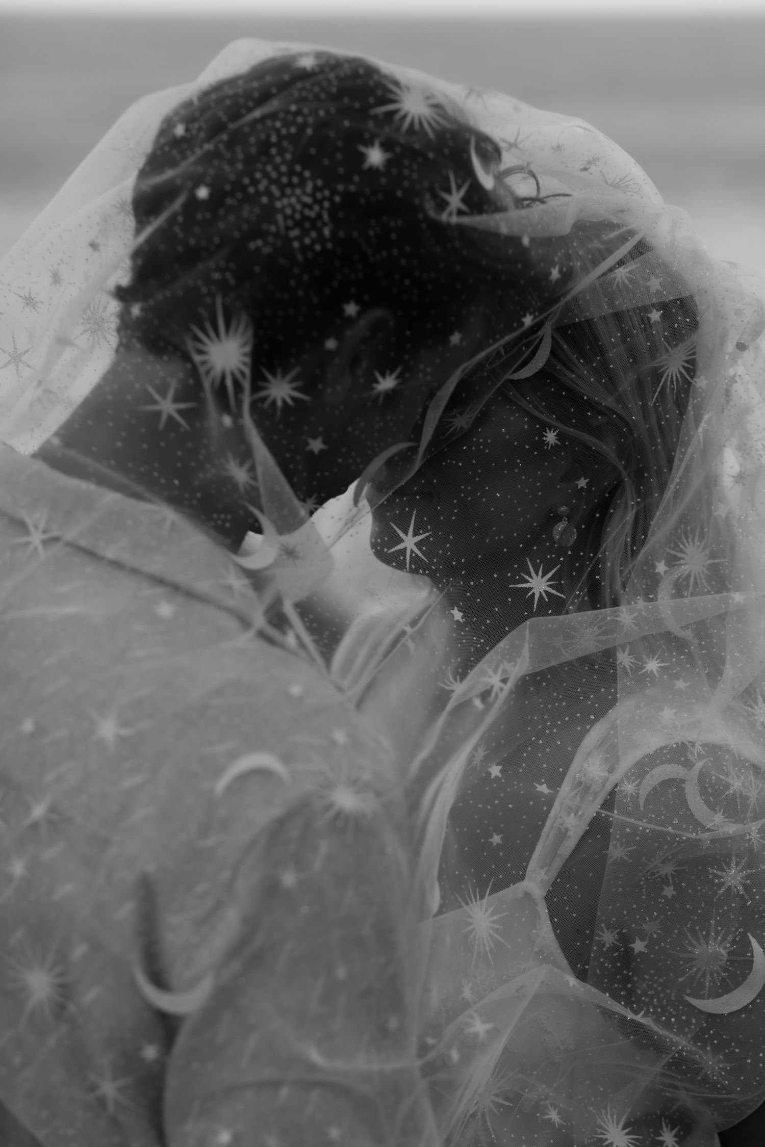 Black and white wedding photo of couple sharing intimate moment under star-embroidered veil.