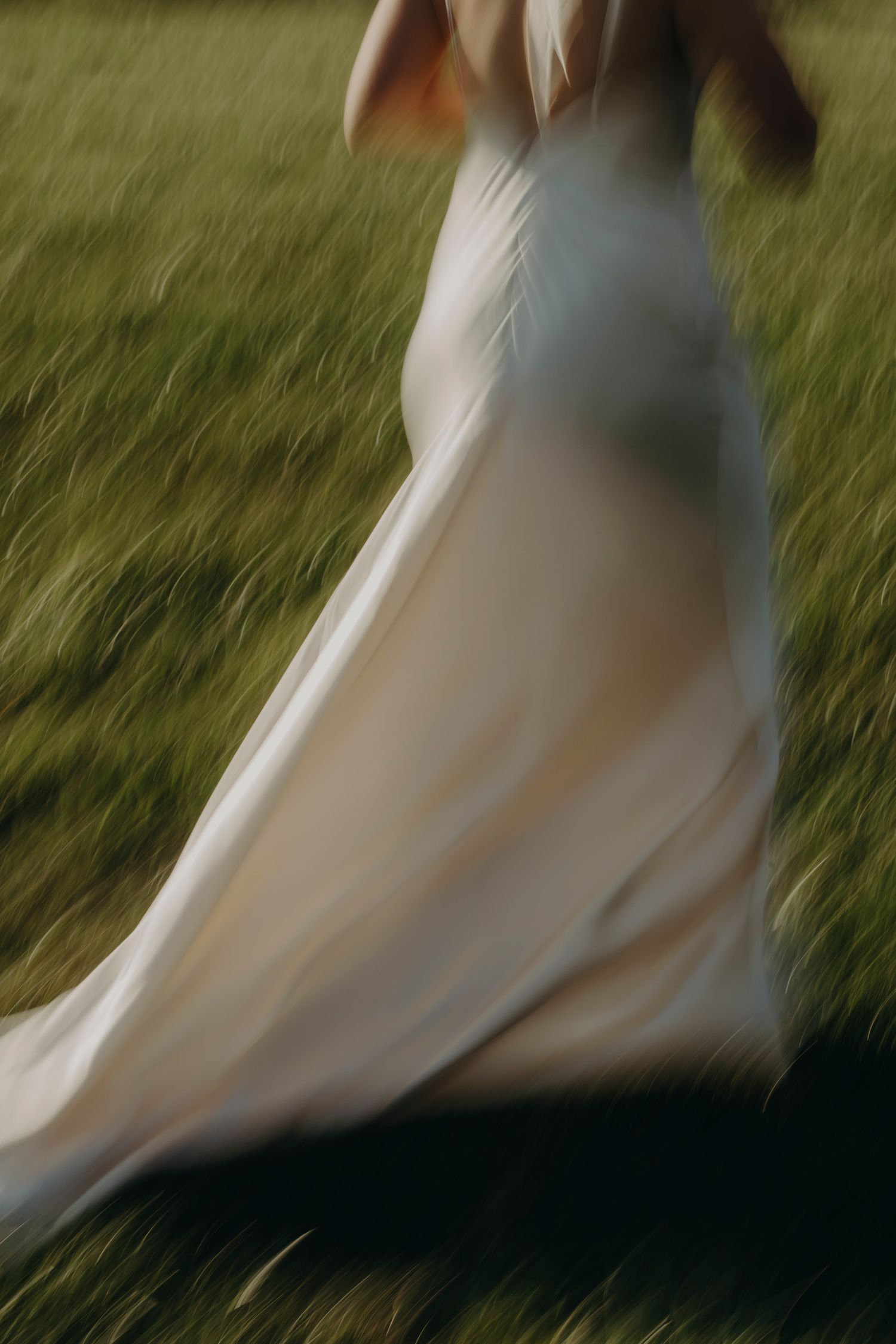 Bride in flowing white wedding gown running through green meadow, motion blur capturing movement.