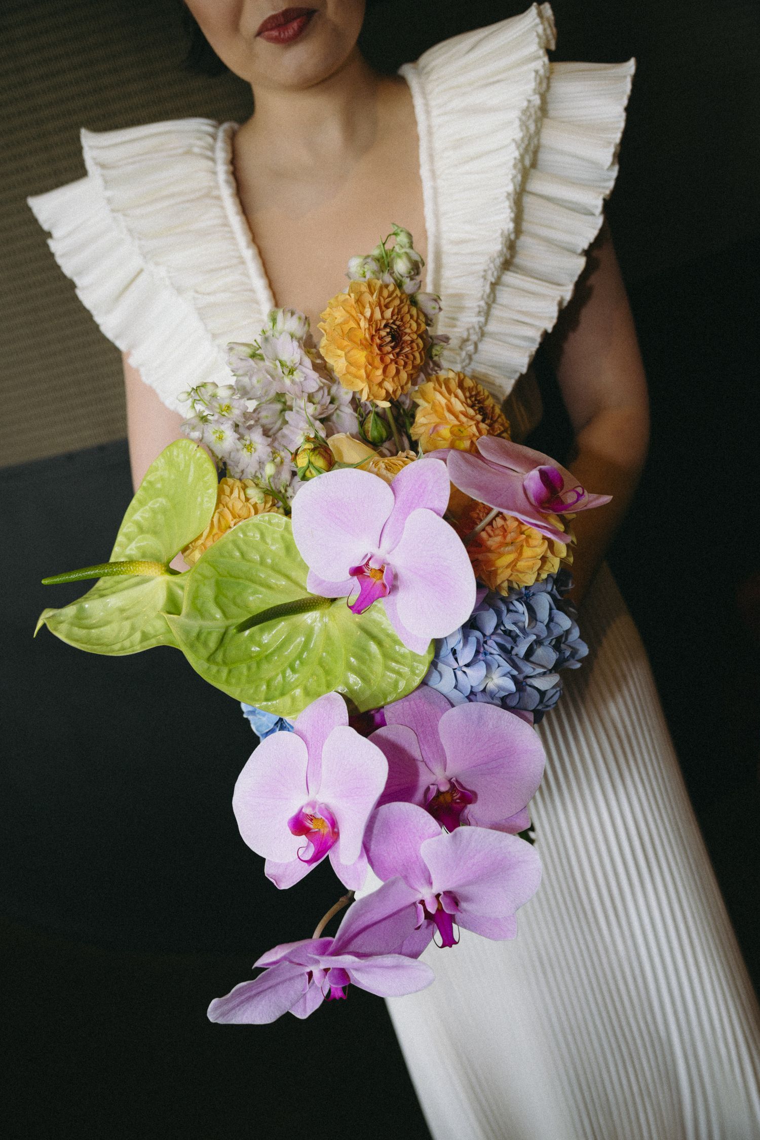 Bride in white ruffled dress holding colorful cascading bouquet with pink orchids and green anthurium.