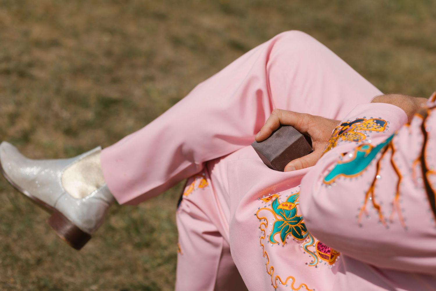 Person sitting on grass wearing pink embroidered pants and white cowboy boots holding a phone.