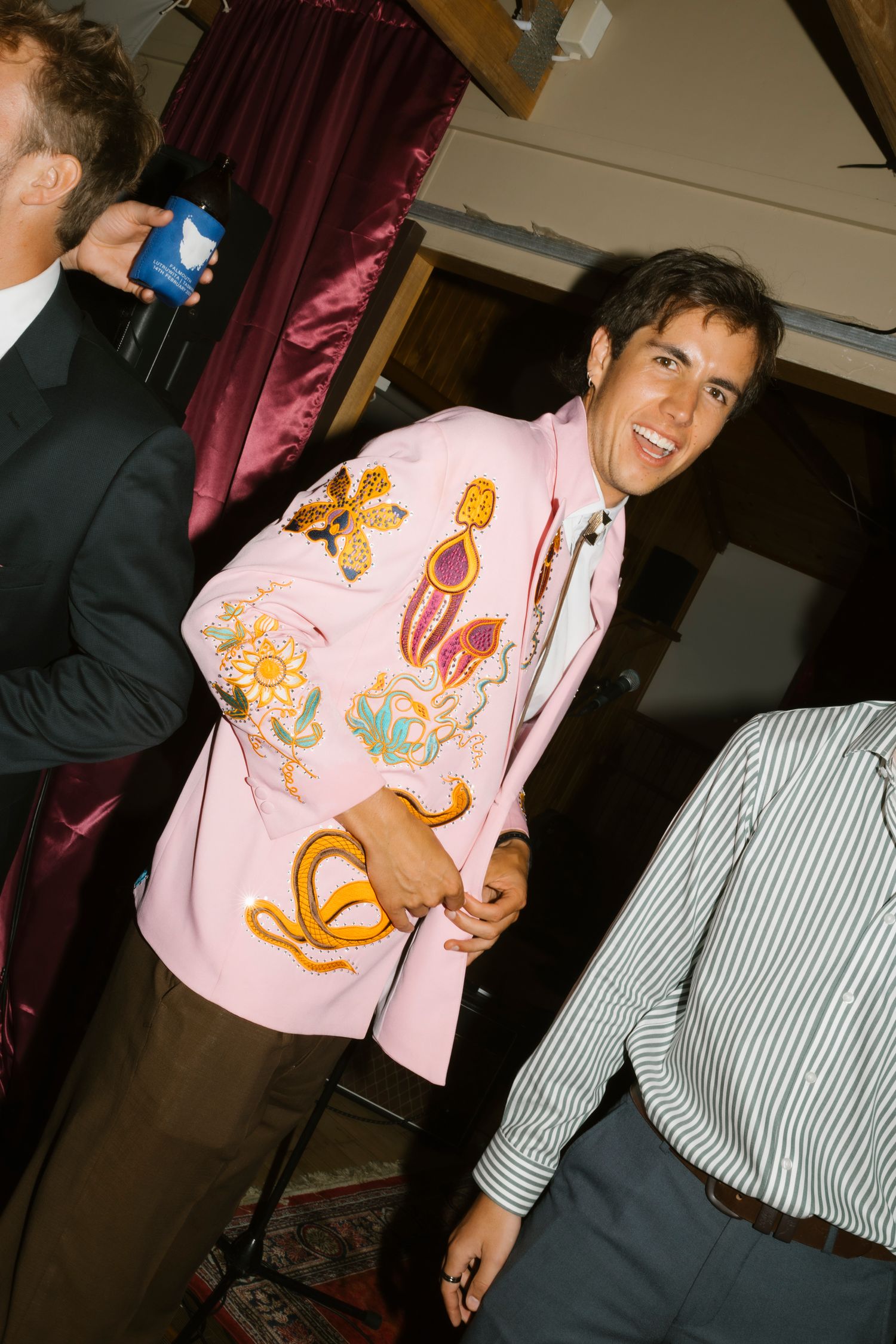 Young man smiling in a pink embroidered blazer at a social gathering with guests in background.