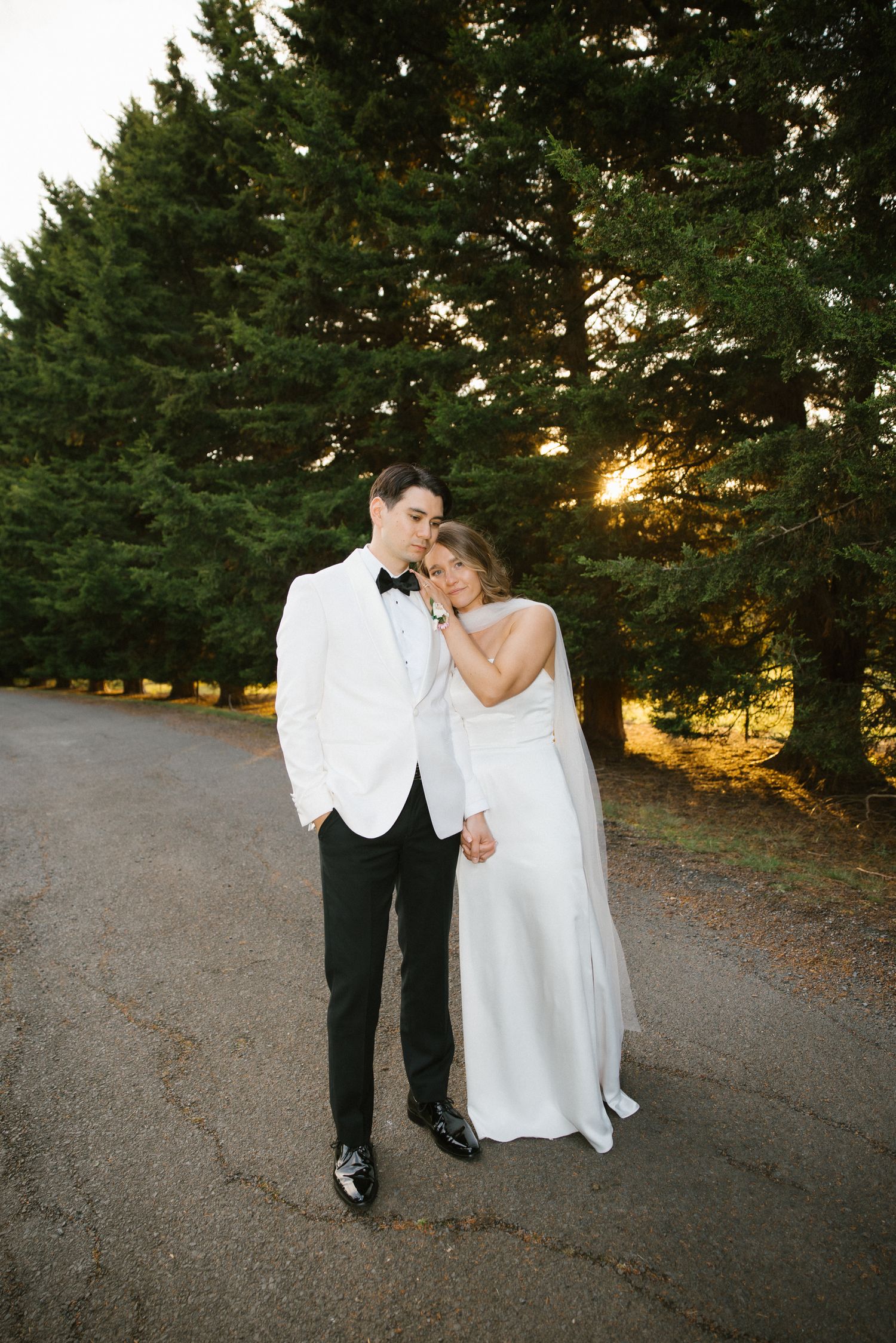 Bride in white gown leaning on groom in white tuxedo jacket among tall pine trees at golden hour.