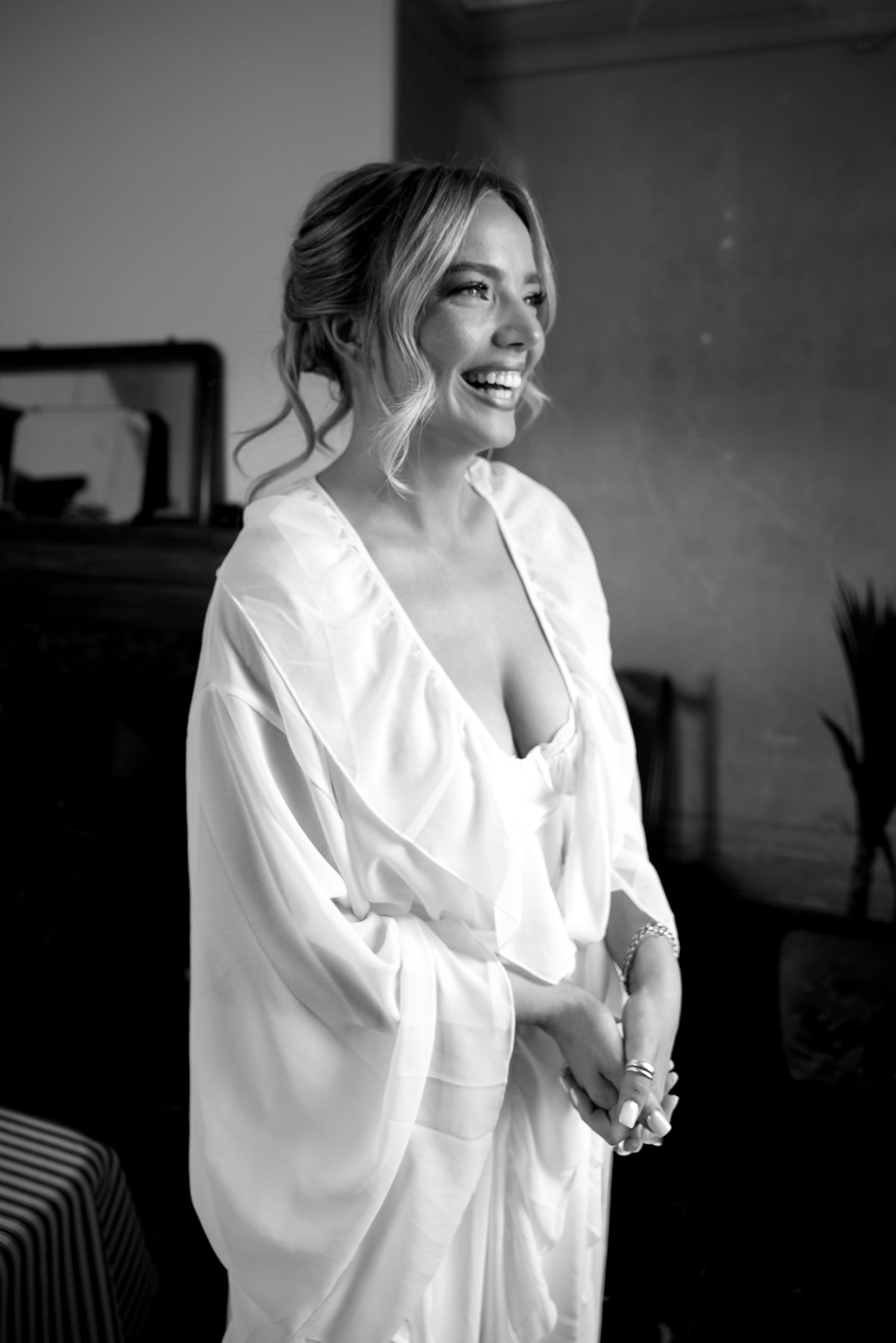 Black and white photo of radiant bride in white robe smiling joyfully during wedding preparations.