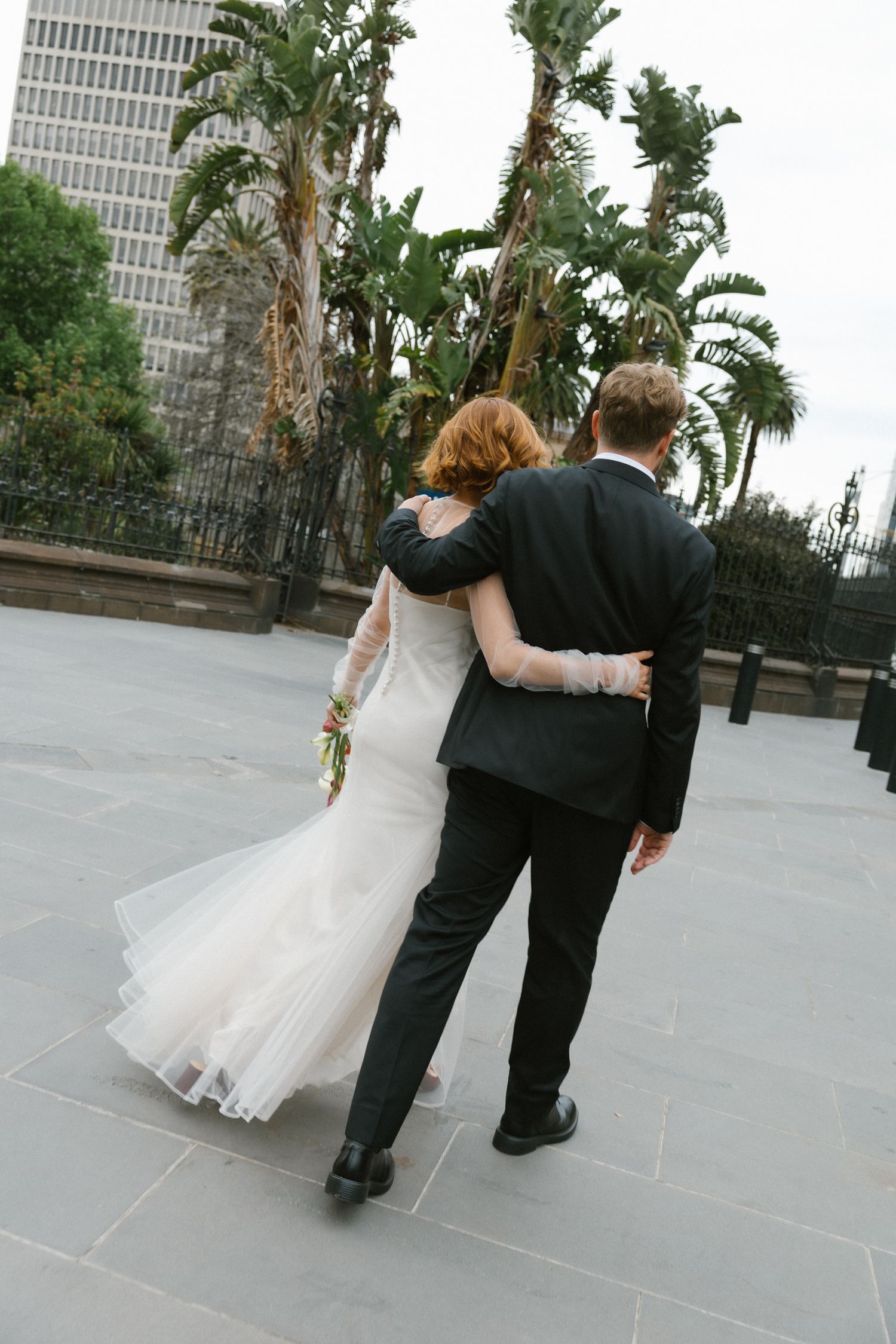 Newlyweds walk arm in arm away from camera past tropical palm trees on paved plaza.