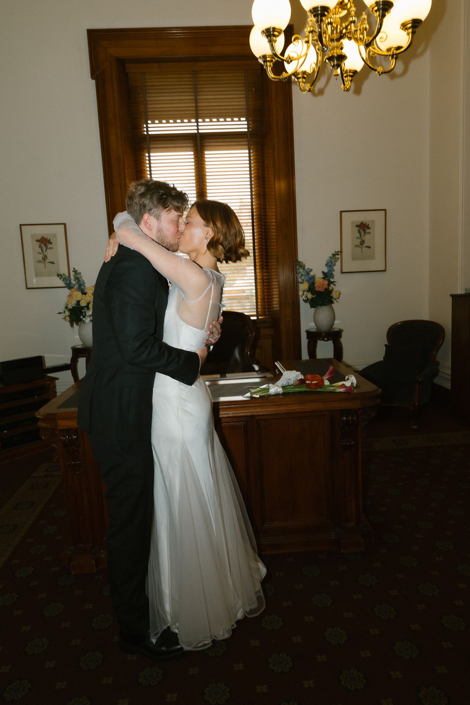 Newlyweds share first kiss in elegant wood-paneled room with chandelier after civil ceremony.