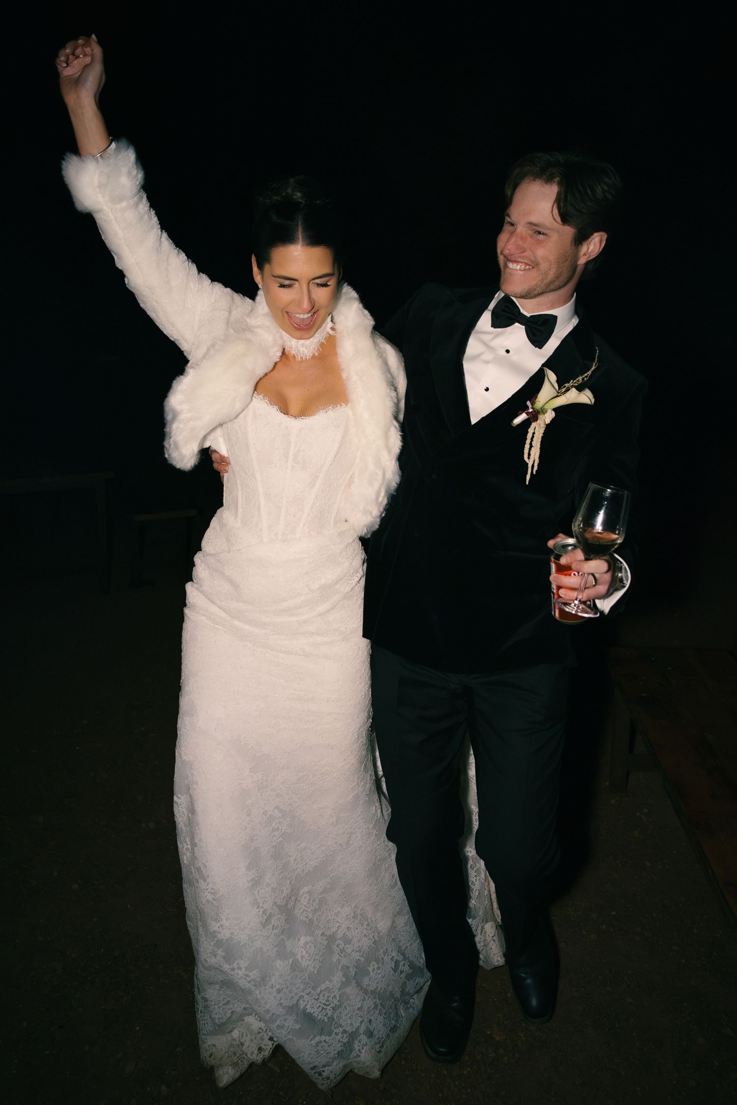 Joyful bride and groom celebrating outdoors at night, bride raising her arm in excitement.