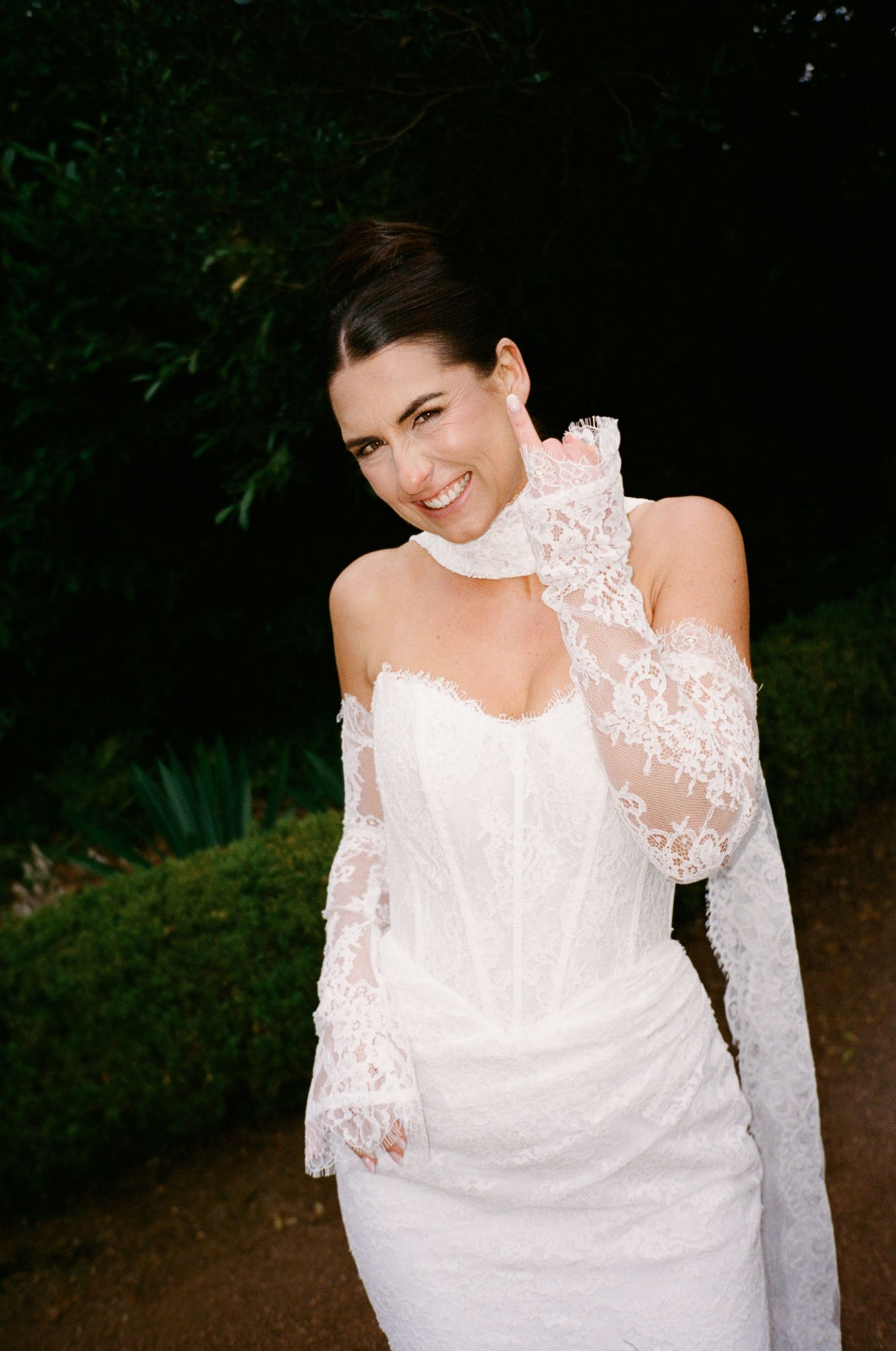 Smiling bride in white lace off-shoulder gown with matching lace gloves poses outdoors against dark green foliage.