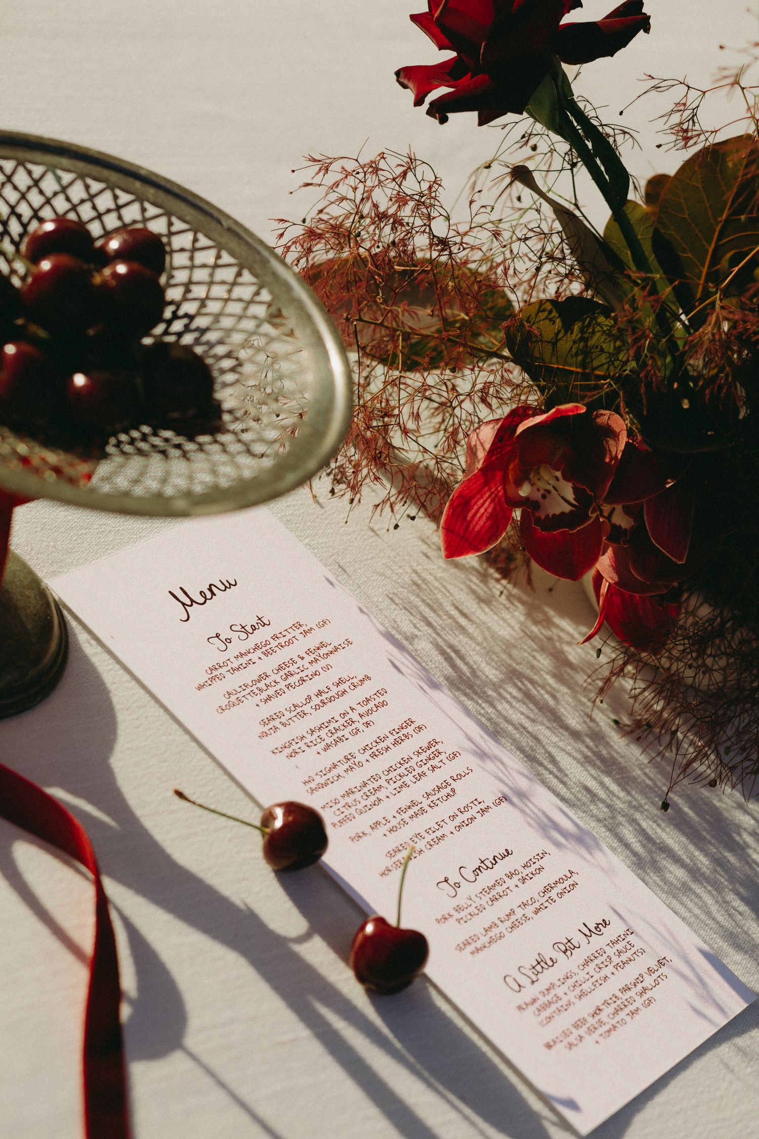 Elegant wedding menu card on white linen with red roses, cherries and ribbon details.
