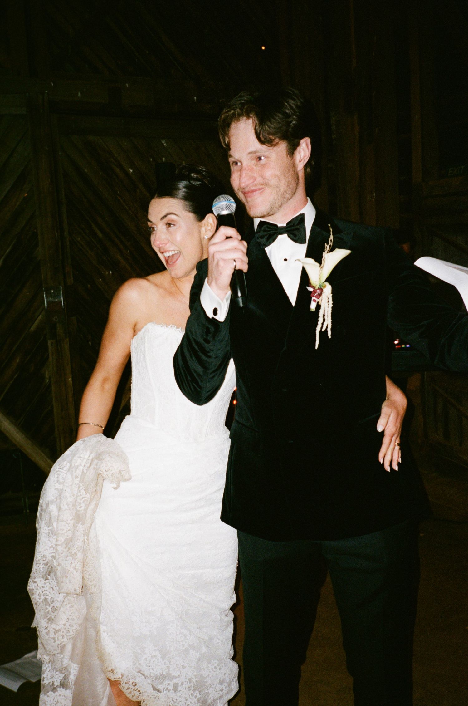 Bride in white strapless gown and groom in black tuxedo sharing a microphone at their wedding reception.