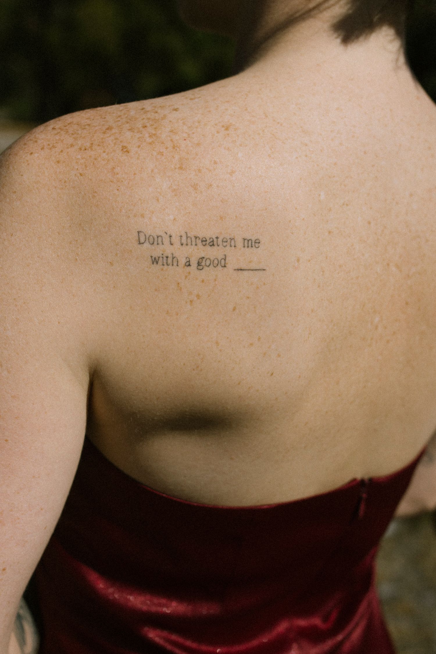 Freckled shoulder tattoo reading Don't threaten me with a good on a woman in red dress.
