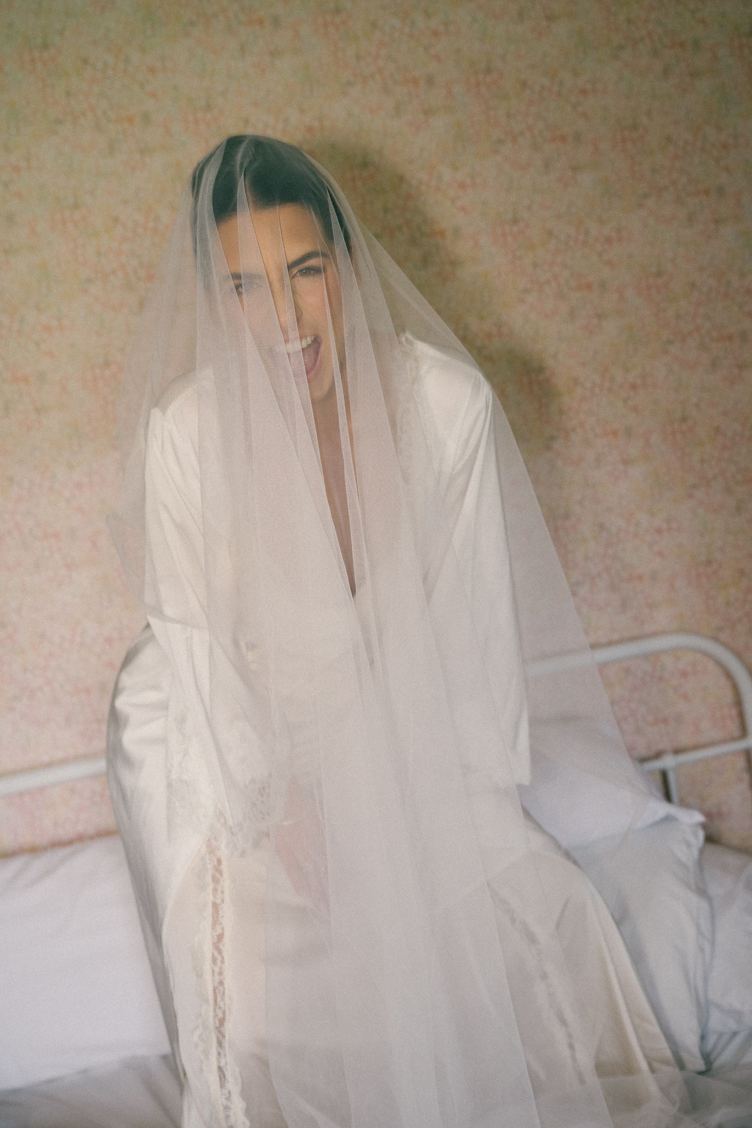 Smiling bride wearing a sheer veil over her face while sitting on a bed in a white robe.