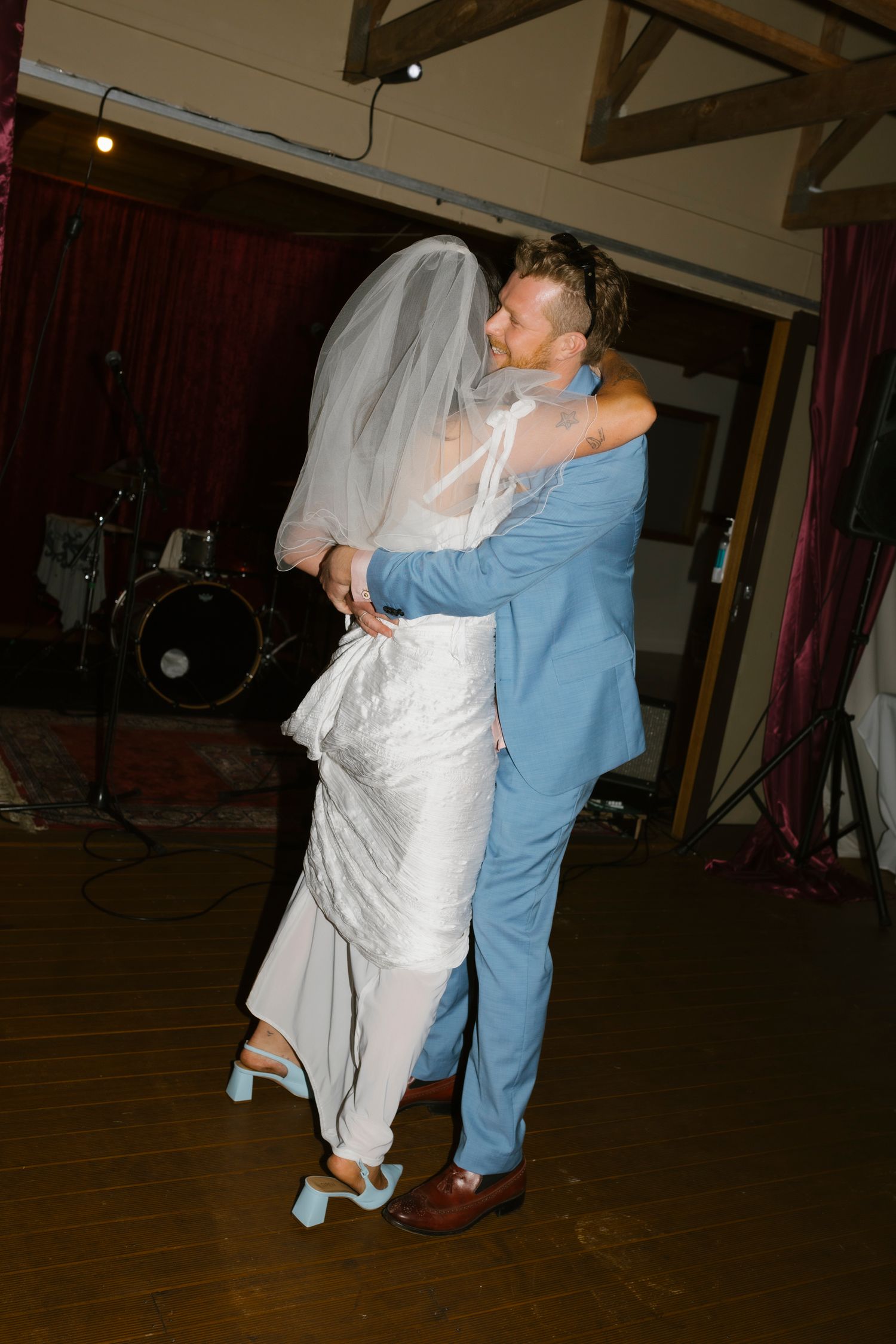 Bride and groom share first dance, embracing on dance floor, bride in white gown and veil, groom in blue suit.