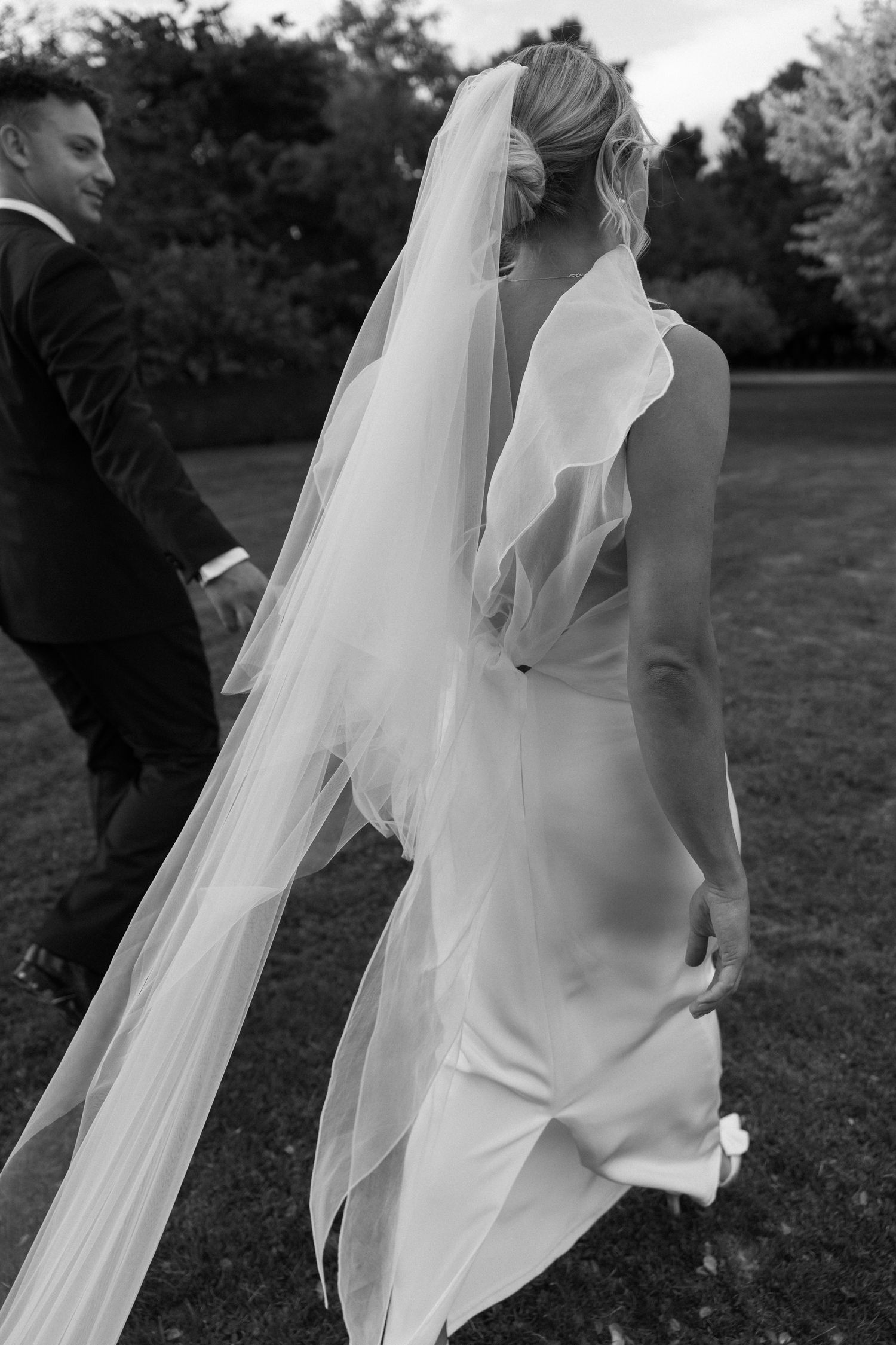 Black and white photo of a bride in a flowing veil walking with her groom in a garden.