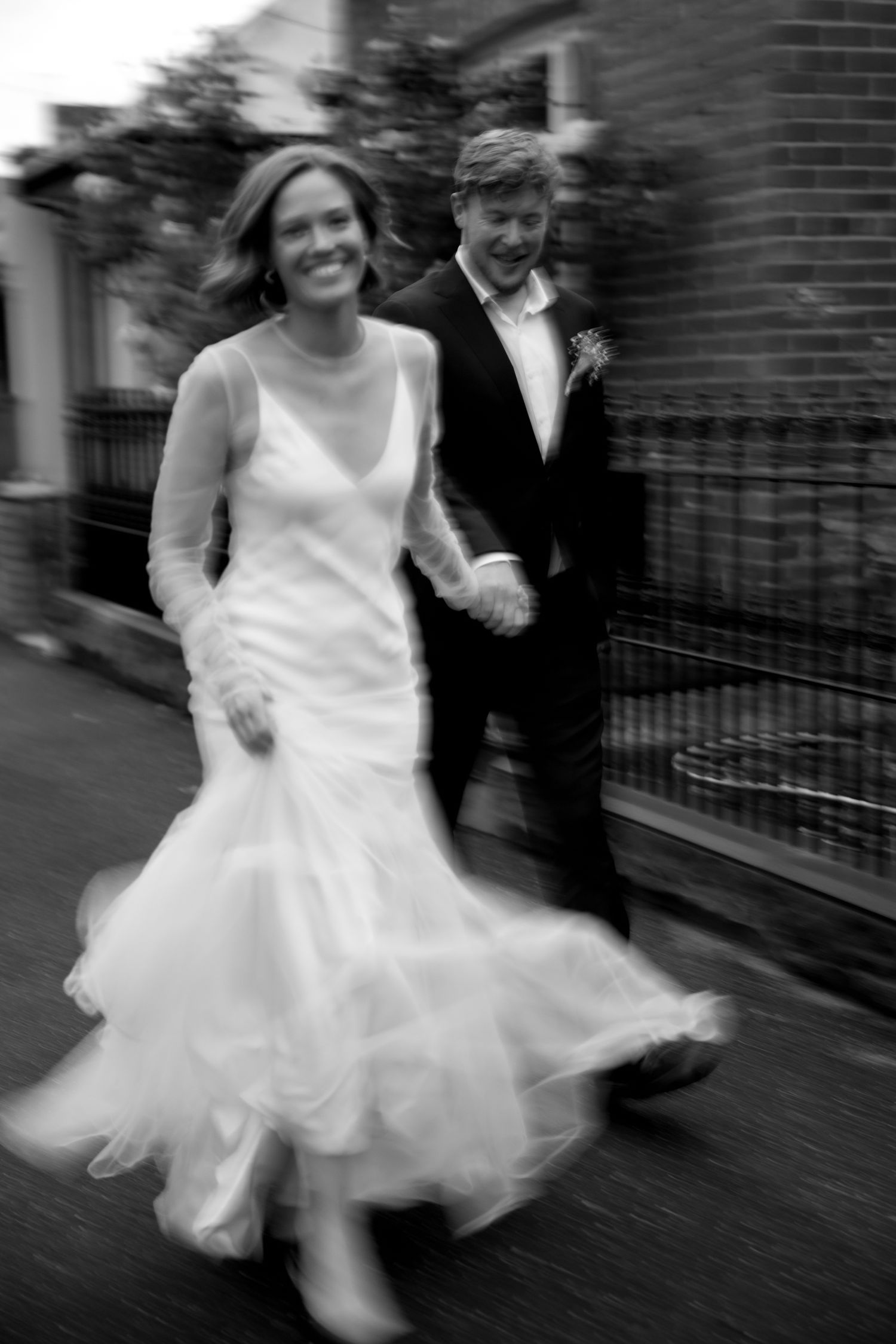 Black and white photo of joyful bride and groom walking hand in hand on a street.
