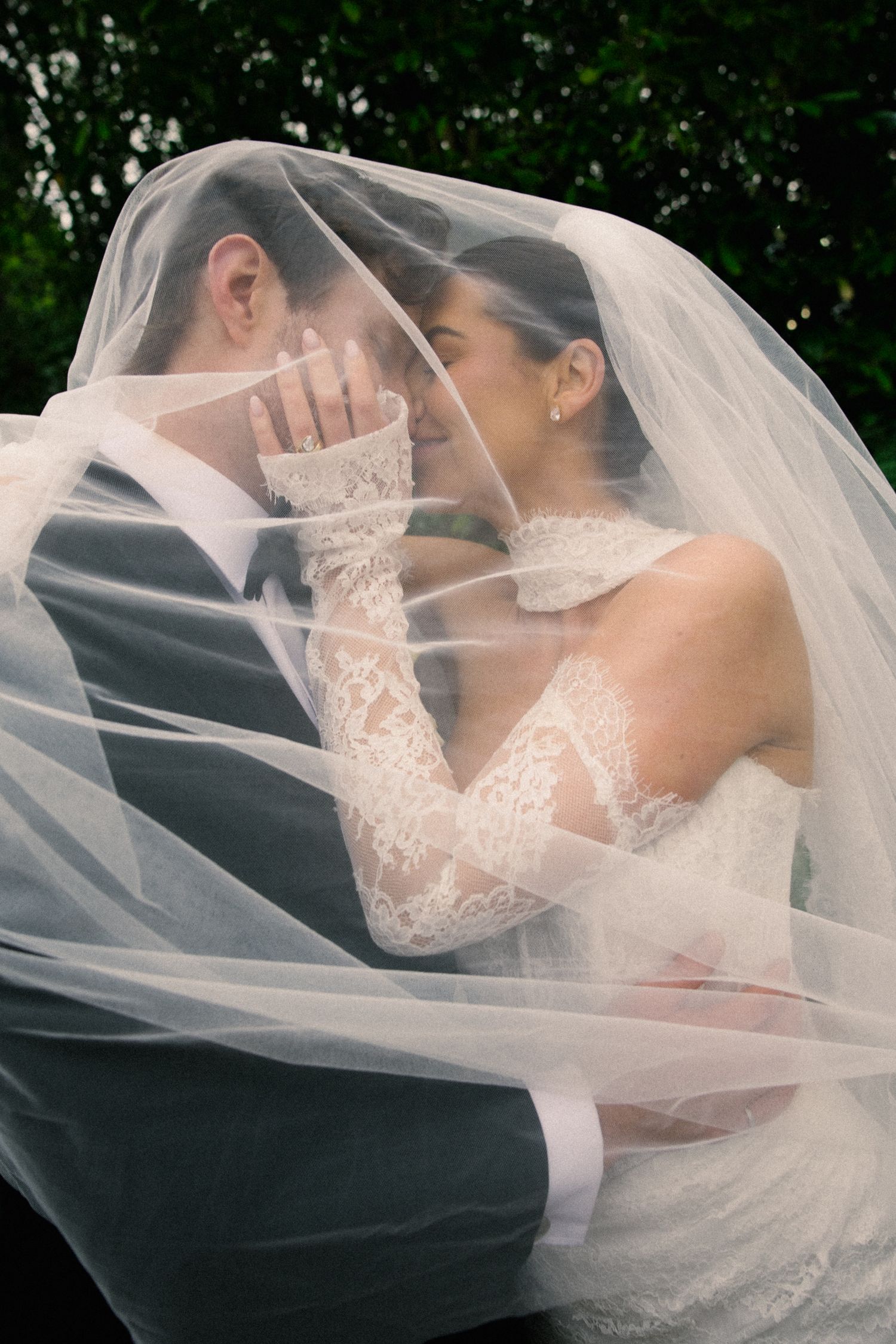Romantic bride and groom sharing an intimate moment under a flowing lace wedding veil outdoors.