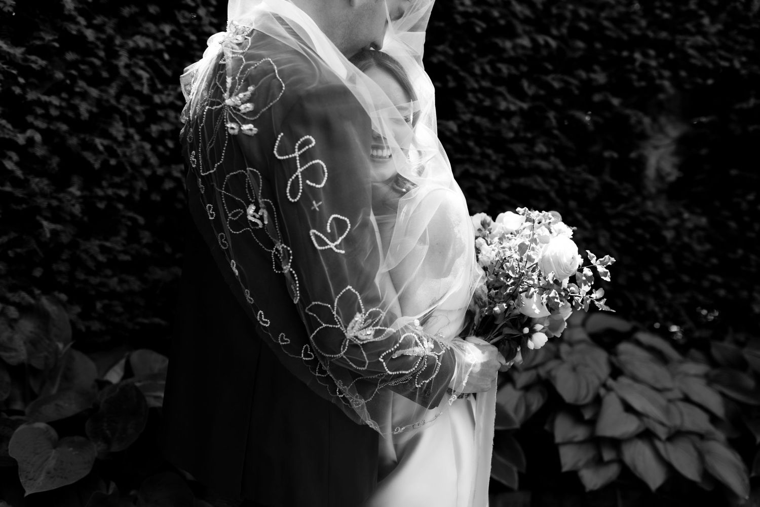Black and white close-up of bride and groom embracing, brides embroidered veil draped over them holding a small bouquet.